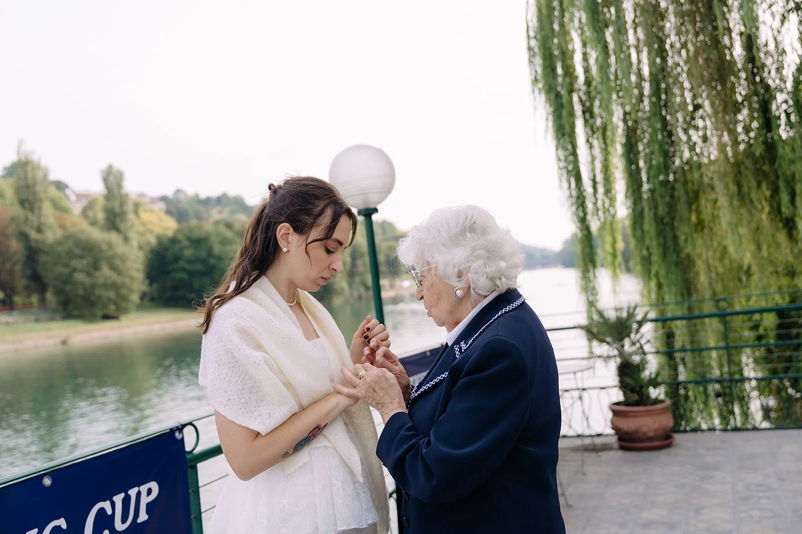 A young woman and an elderly woman holding hands by a river, with trees and a cloudy sky in the background.