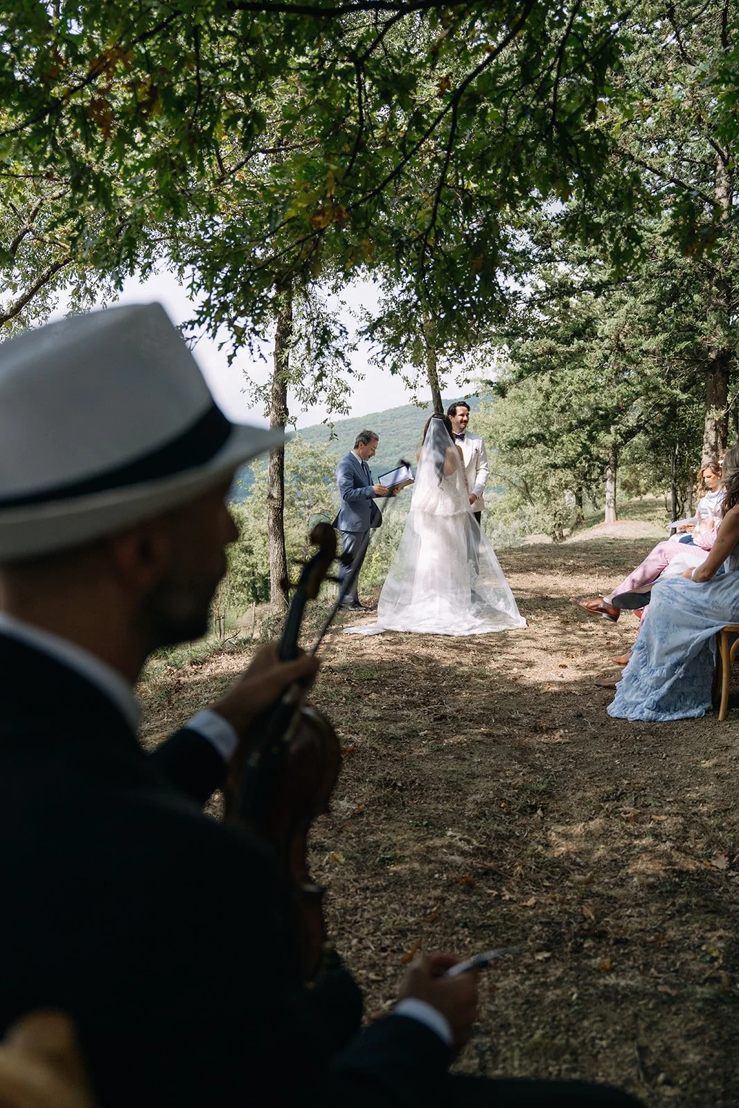 An outdoor wedding ceremony with a bride and groom under trees, officiant reading from a book, guests seated on chairs, musician in foreground playing violin.