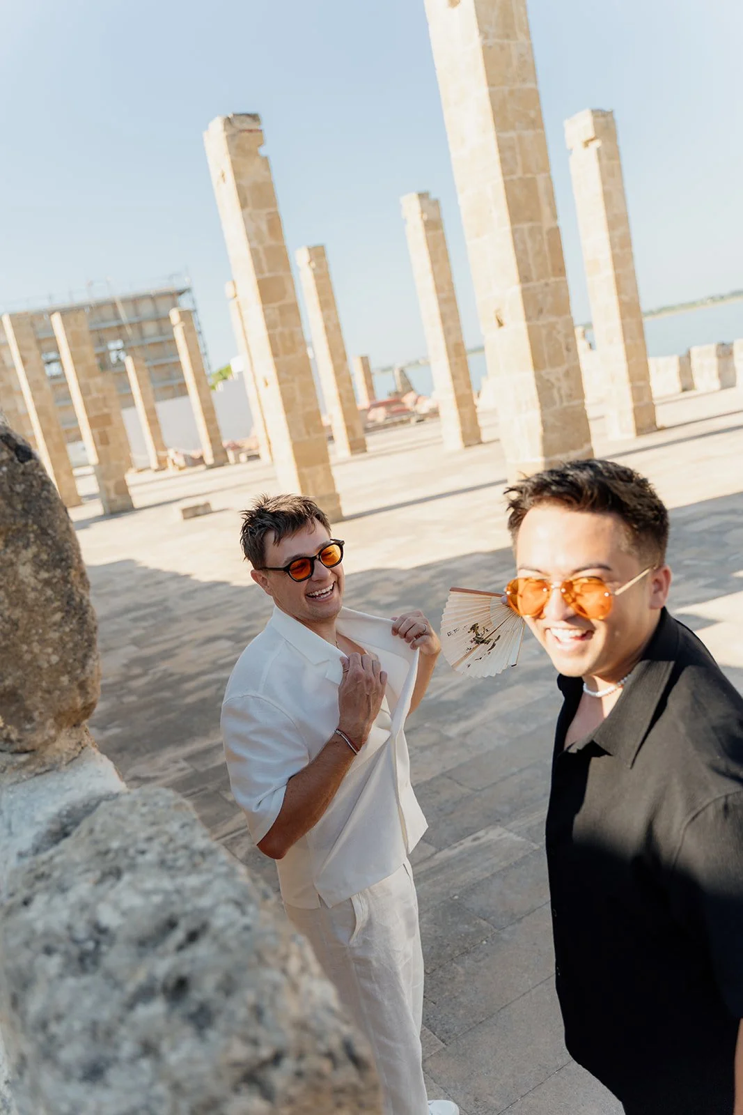 Two men smiling and wearing sunglasses, one with an umbrella, on a sunny day near historical ruins.