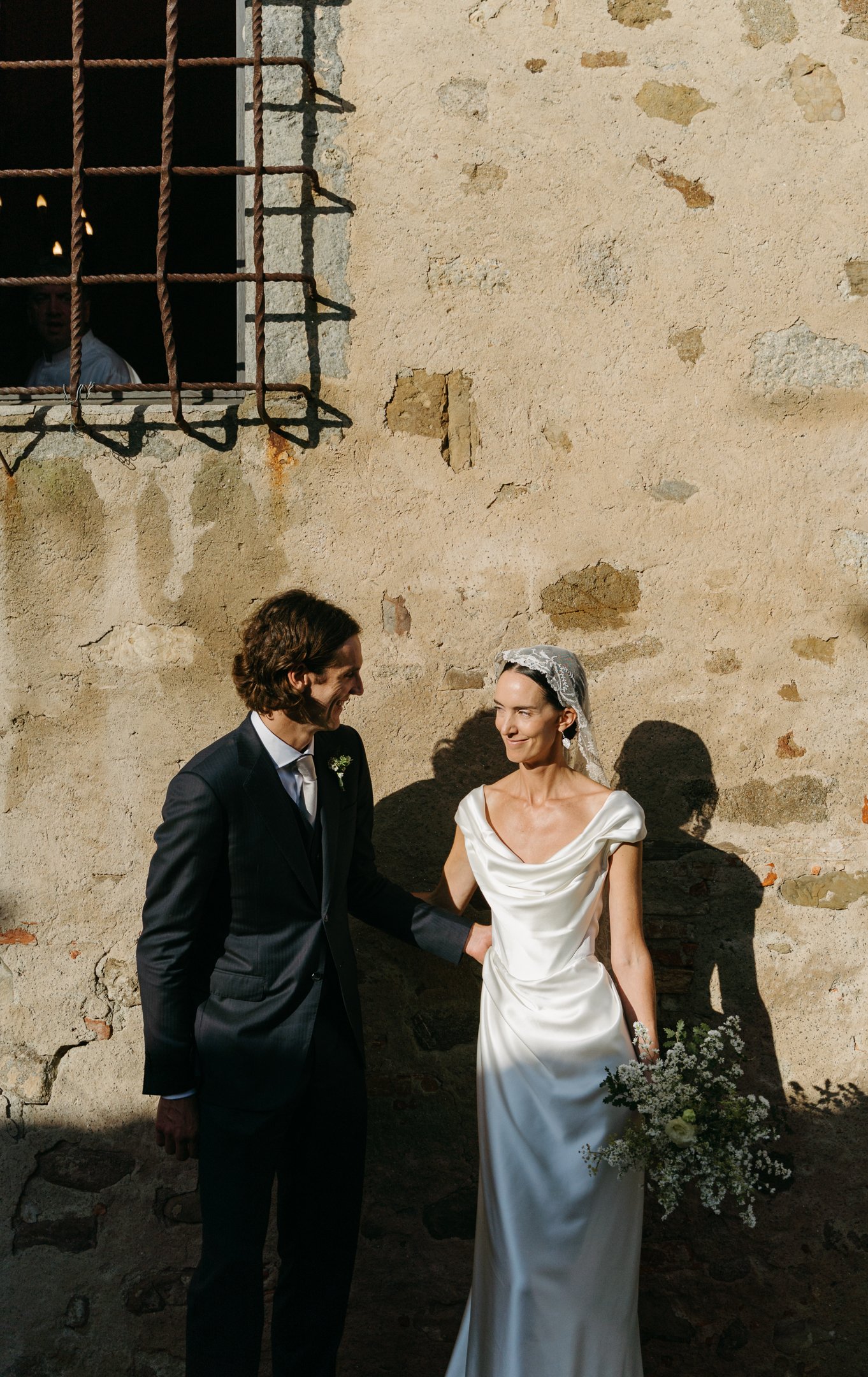 A bride and groom are standing outside against a weathered stone wall, smiling at each other. The bride is holding a bouquet of flowers and wearing a white wedding dress with a lace headpiece. The groom is dressed in a dark suit.