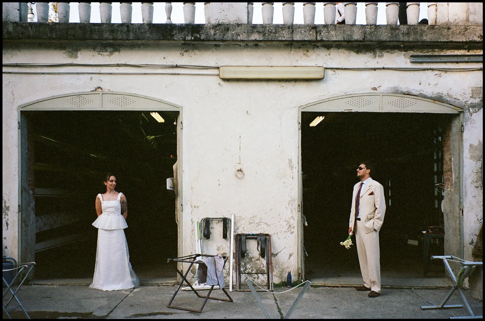 A woman in a white dress and a man in a white suit are standing apart in front of two large open garages with dark interiors. The woman is on the left, looking at the man, who is on the right, holding a small bouquet of flowers. There are chairs, a s