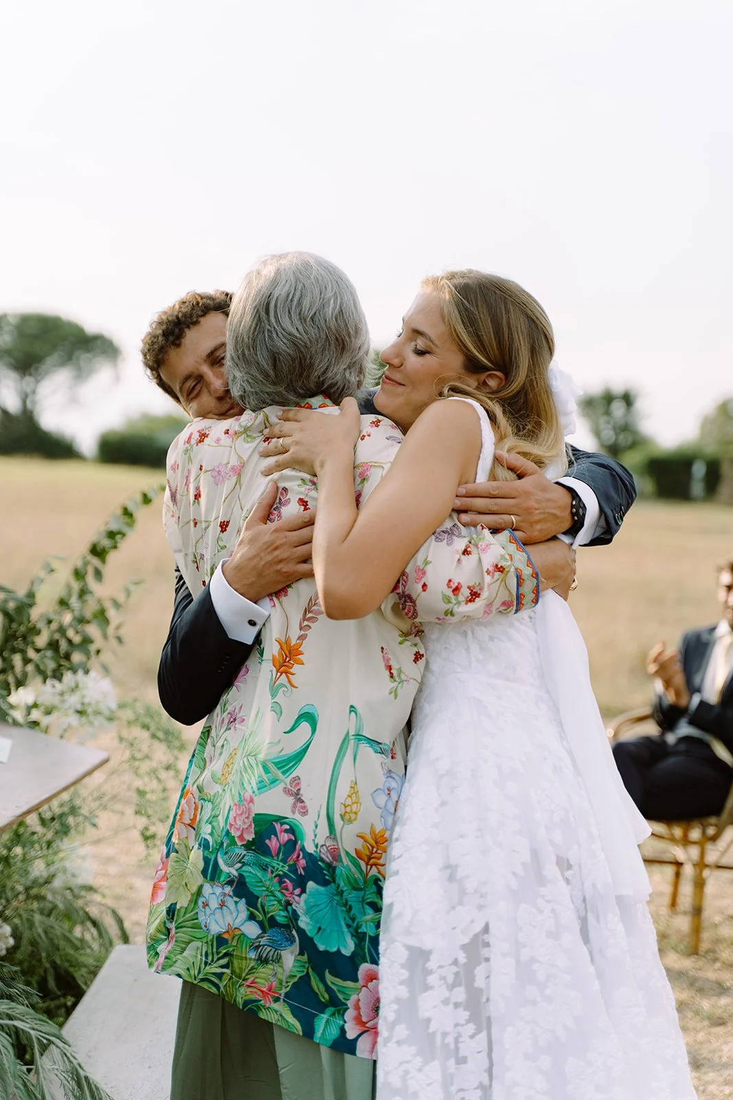 A bride and groom embracing an elderly woman outdoors during a wedding ceremony.