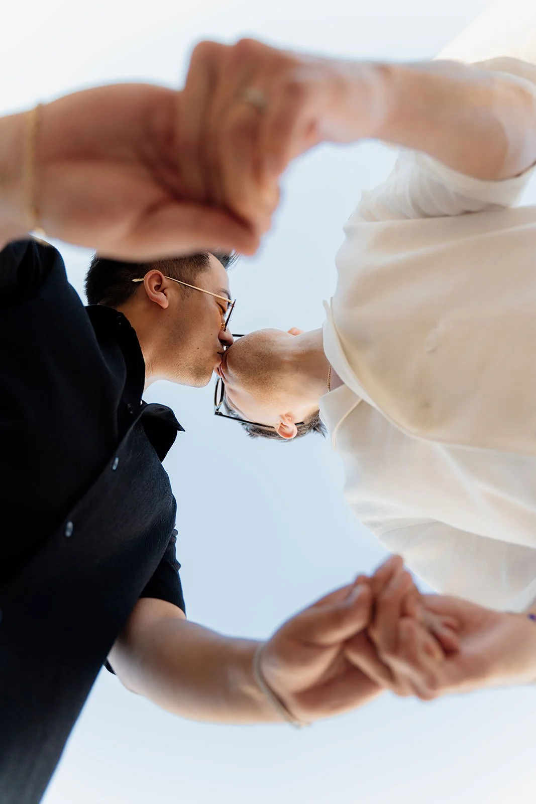 Two men kissing while holding hands, taken from below against a clear sky.