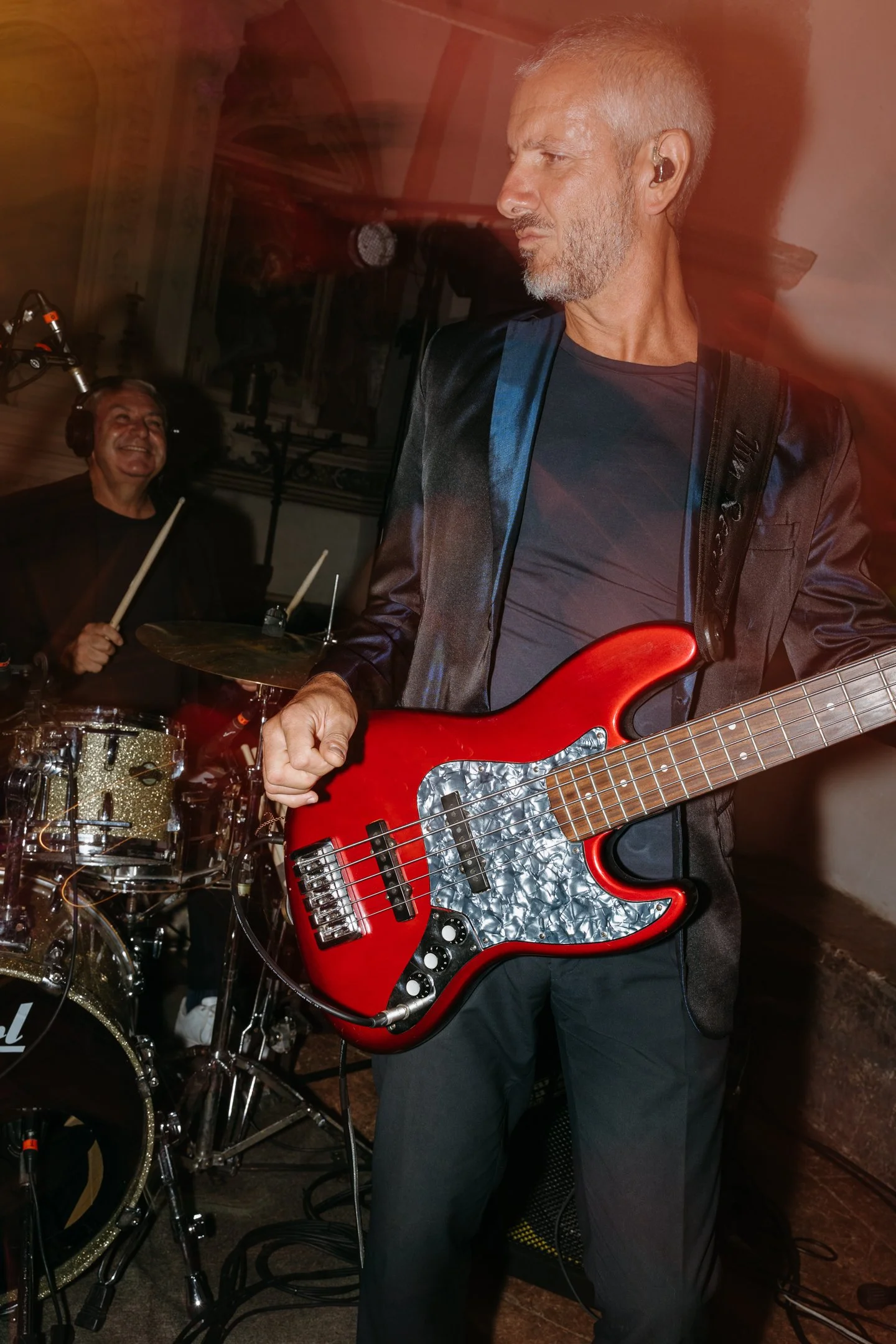 Two musicians performing on stage, one playing the drums and the other playing a red electric bass guitar.