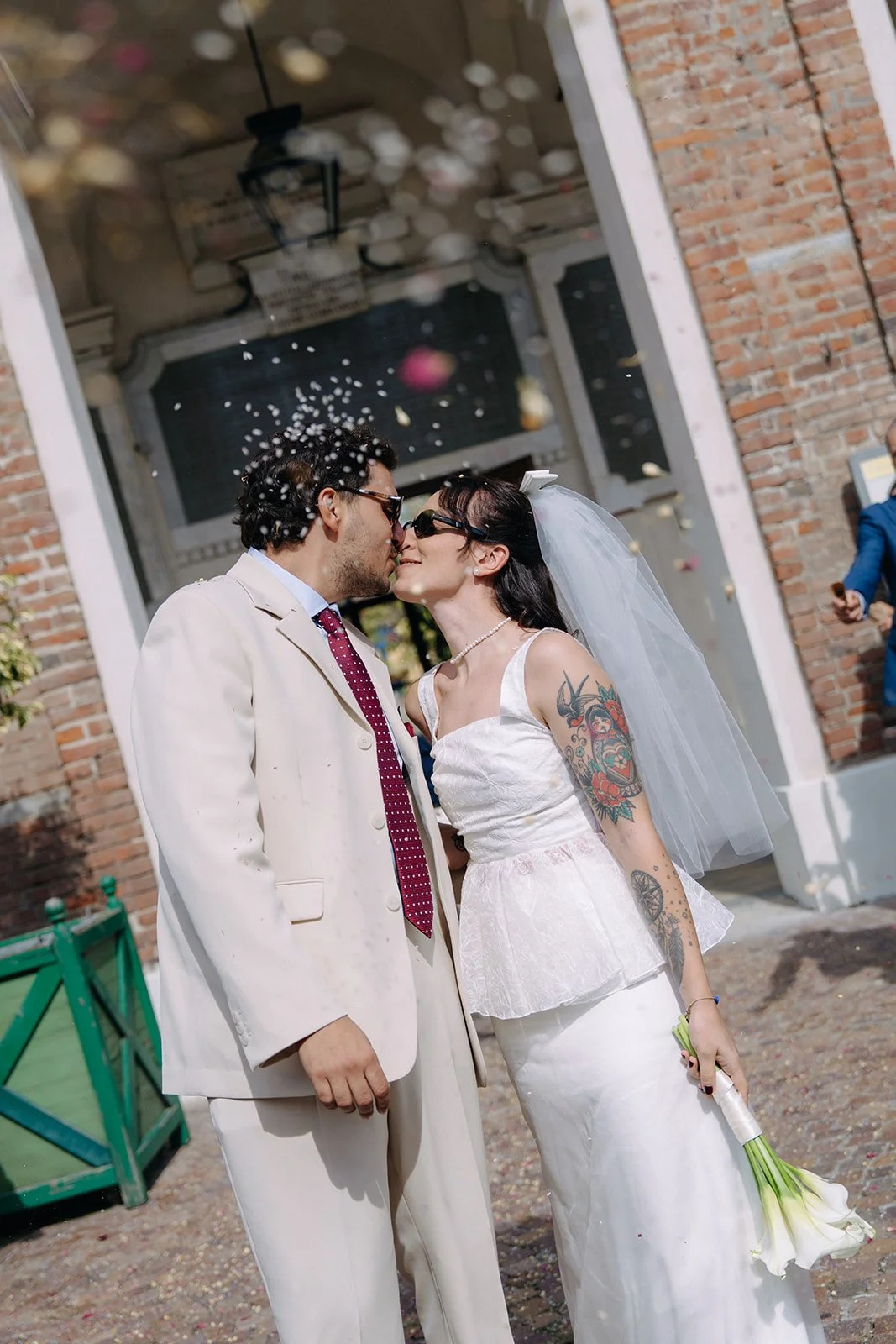 A couple dressed in wedding attire kissing outside, with rose petals falling around them. The groom wears a light-colored suit with a red polka dot tie, and the bride wears a white wedding dress with tattoos on her arms, holding a bouquet of white ca