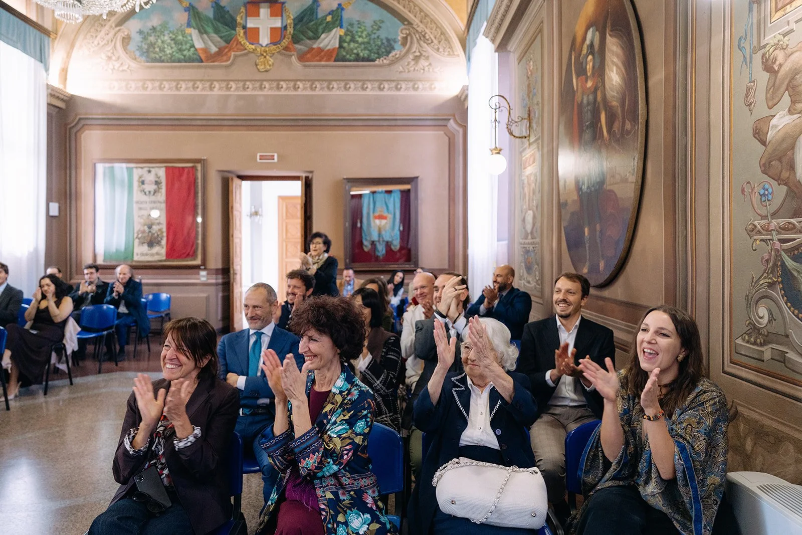 People sitting and clapping in a decorated room with Italian flags and painted murals, attending a formal event.