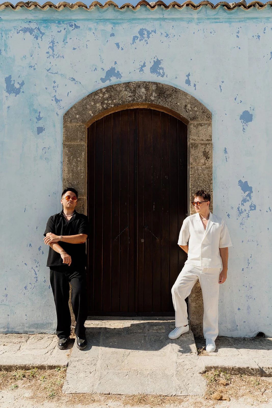 Two people standing in front of a large dark wooden door set into a light blue, weathered wall. The person on the left wears black clothing and sunglasses, leaning against the wall with crossed arms. The person on the right wears a white outfit and s