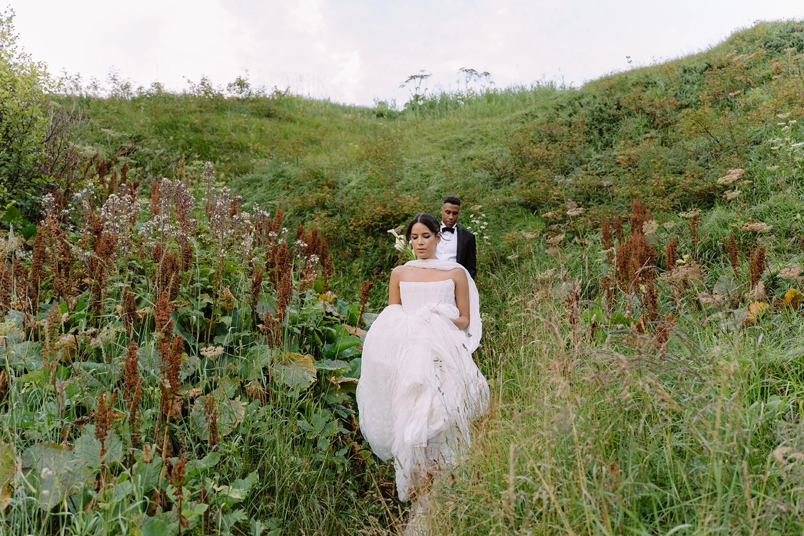 Bride and groom walking through a grassy, hilly landscape with lush greenery and wildflowers.