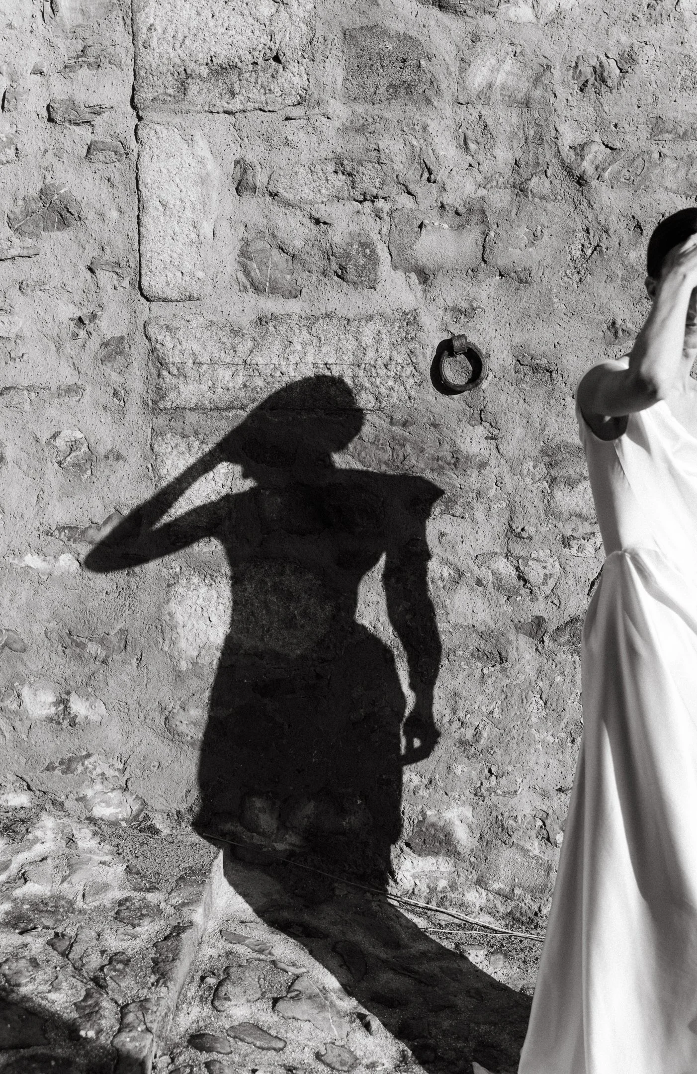 Shadow of a woman holding a hat on a textured stone wall, with part of a woman in a white dress visible on the right.