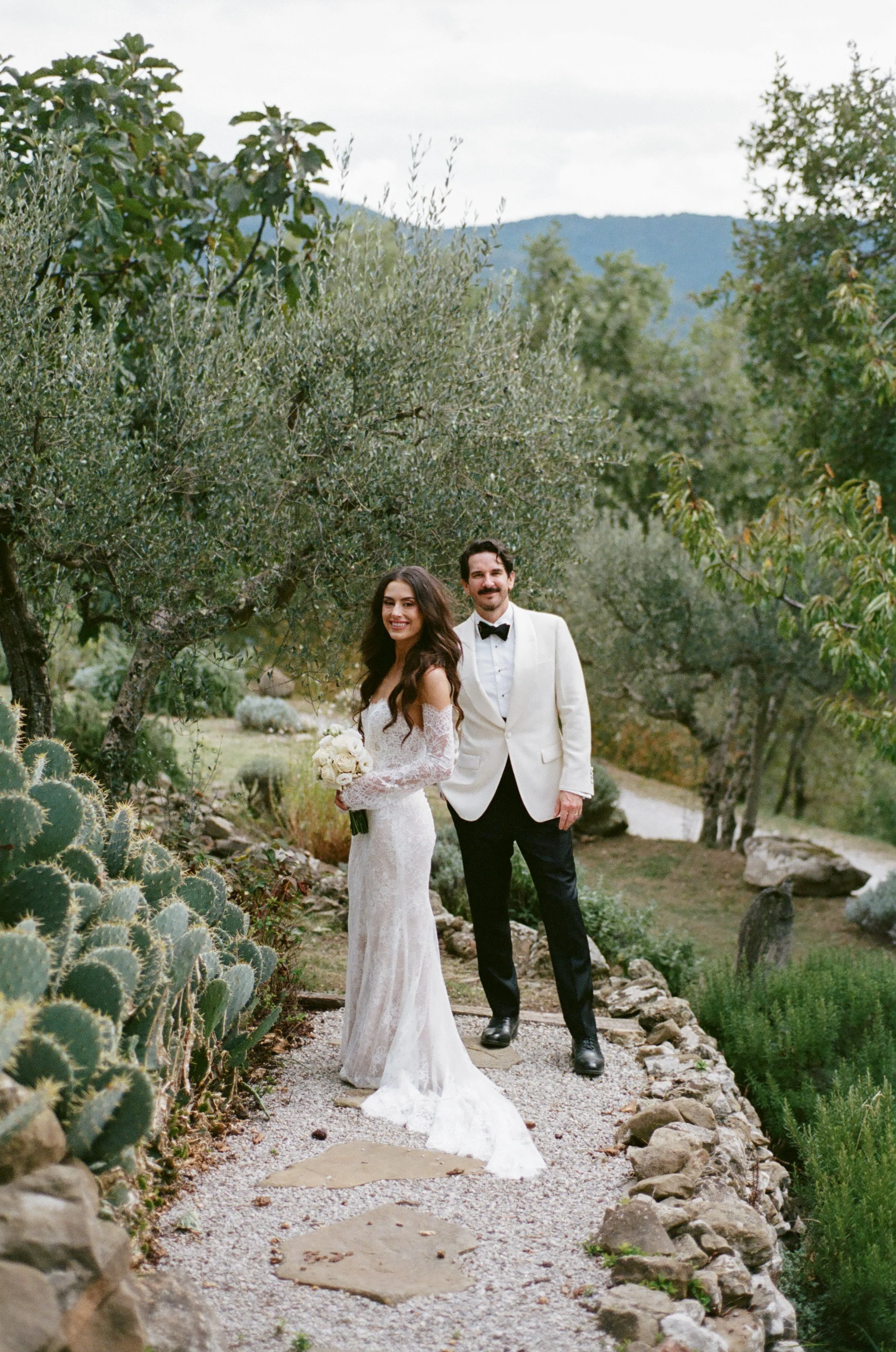 A bride and groom standing on a garden pathway, surrounded by trees and plants, with mountains in the background. The bride is wearing a white lace wedding dress and holding a bouquet, and the groom is dressed in a white tuxedo jacket and black pants