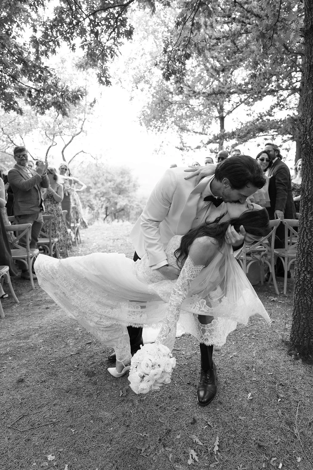 A black-and-white photo of a wedding scene with a bride and groom. The groom is dipping the bride and kissing her forehead. The bride is holding a bouquet of flowers, and her wedding dress is lace. Guests are standing and sitting around in an outdoor