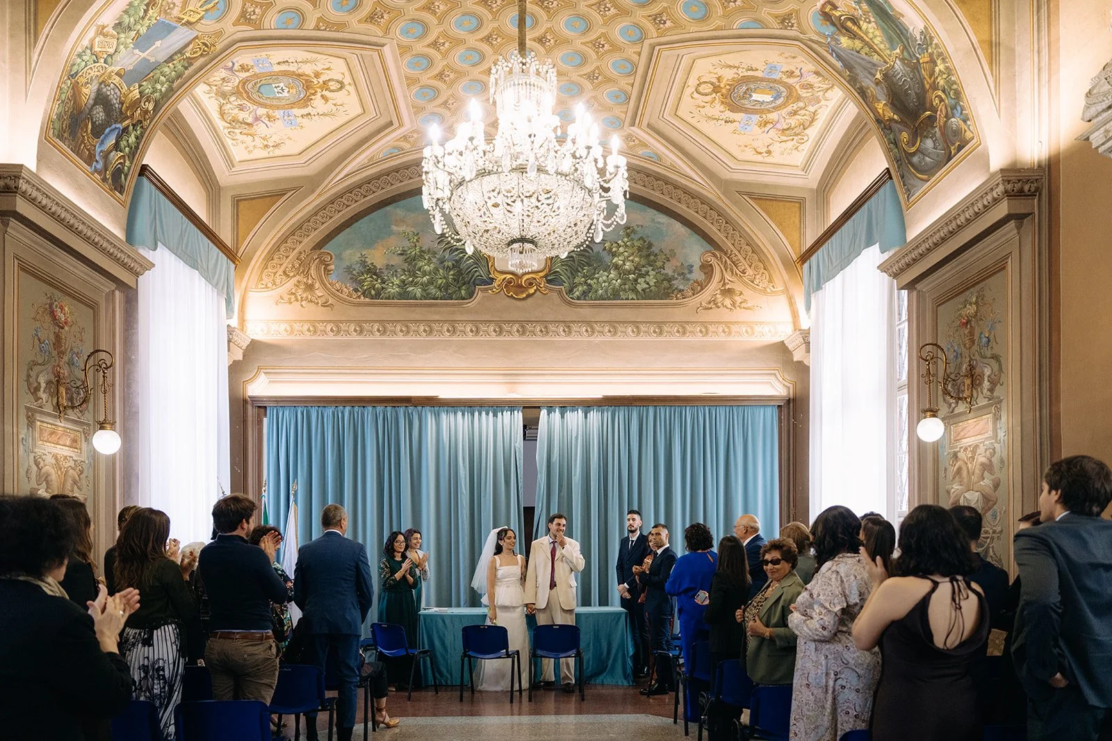 A wedding ceremony in a decorated, ornate hall with a chandelier, large windows with curtains, and a group of people standing and clapping around the couple.