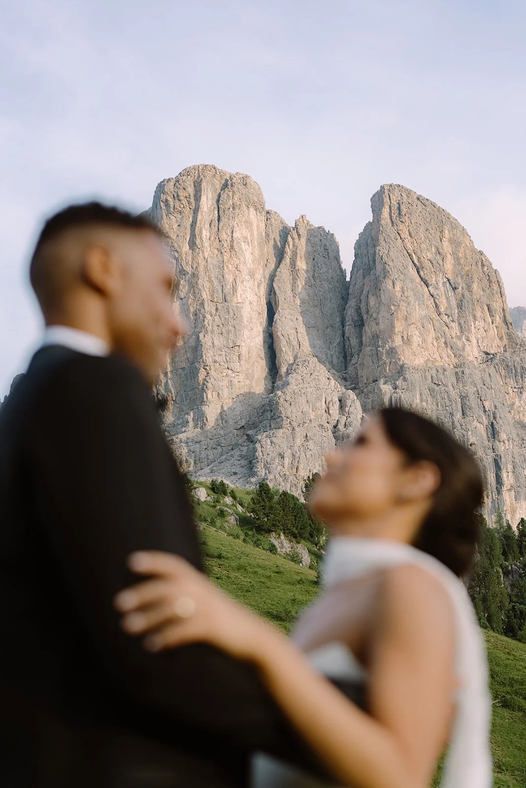 A blurred couple, a man in a suit and a woman in a dress, embracing outdoors with mountains and green hills in the background.