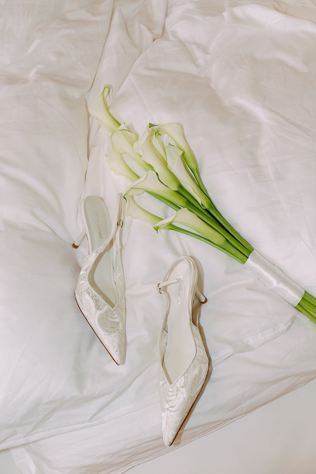 A pair of white satin high-heeled shoes with lace embroidery and pointed toes, and a bouquet of white calla lilies, all arranged on a white fabric background.