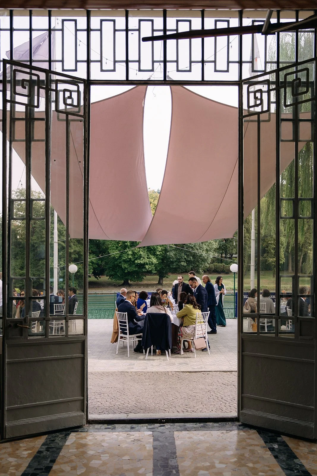 People dining outdoors on a terrace, viewed through an open door with geometric metalwork, with a large pink fabric canopy overhead and greenery in the background.