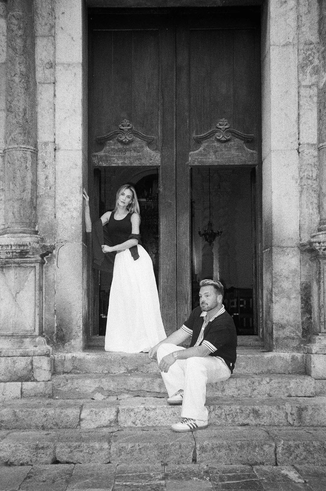 A woman and a man with light skin posing in front of large wooden doors on stone steps; woman standing in the doorway wearing a sleeveless top and long skirt, holding a jacket, man sitting on the steps wearing a polo shirt and white pants.
