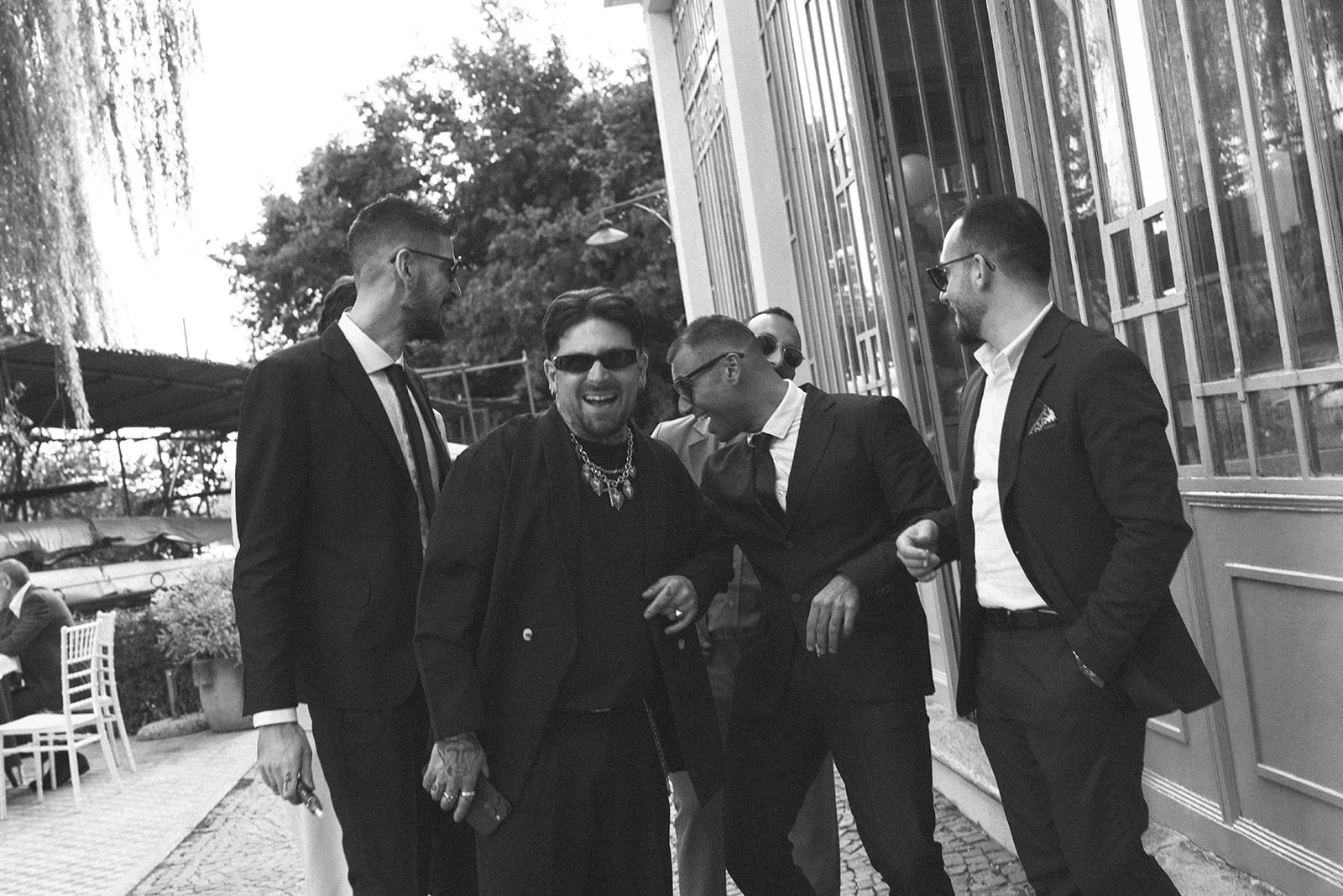 A group of six well-dressed men laughing and talking outside a building, with outdoor furniture and trees in the background.