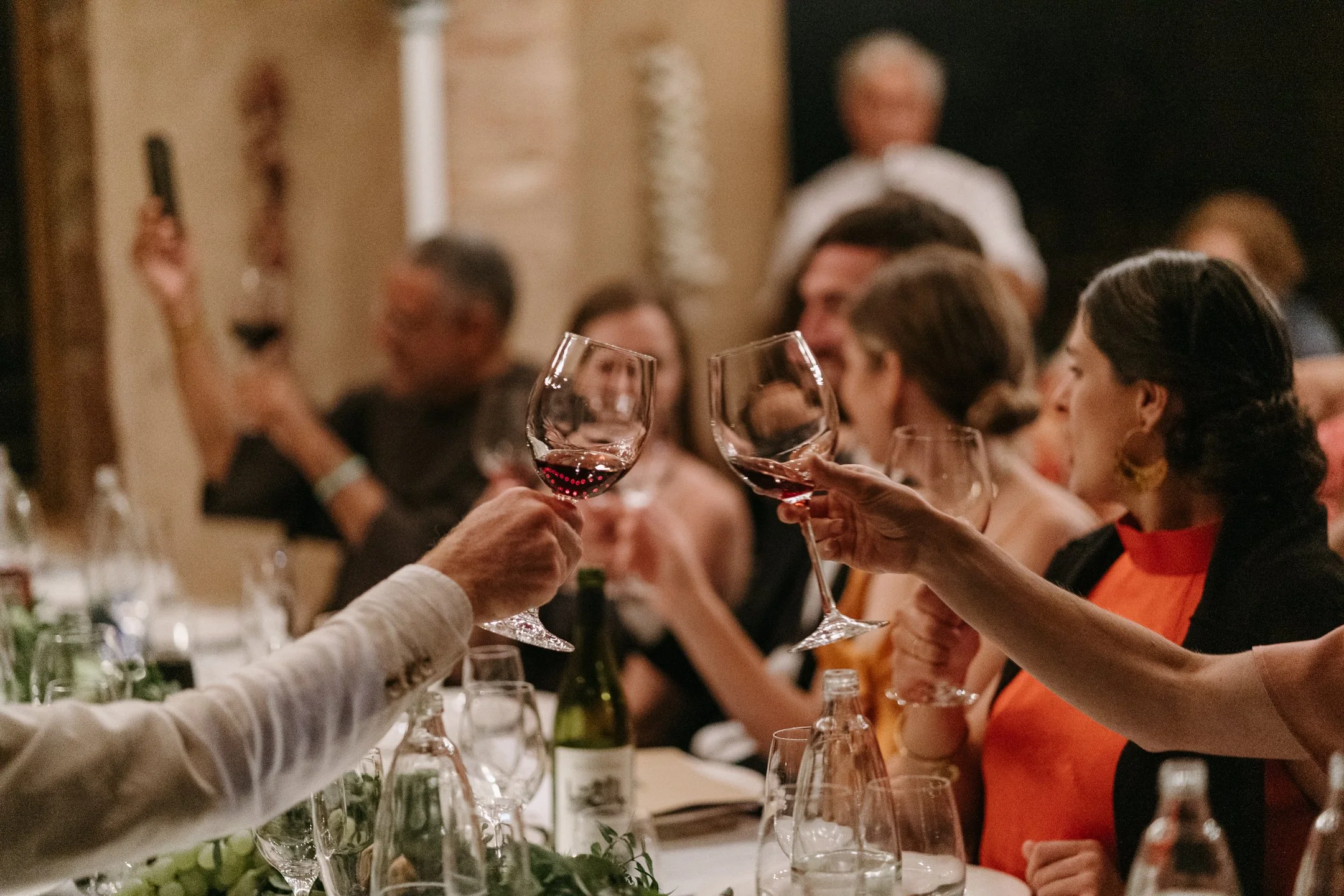 Group of people clinking wine glasses at a dinner party, with a table set with bottles, glasses, and decorative greenery.