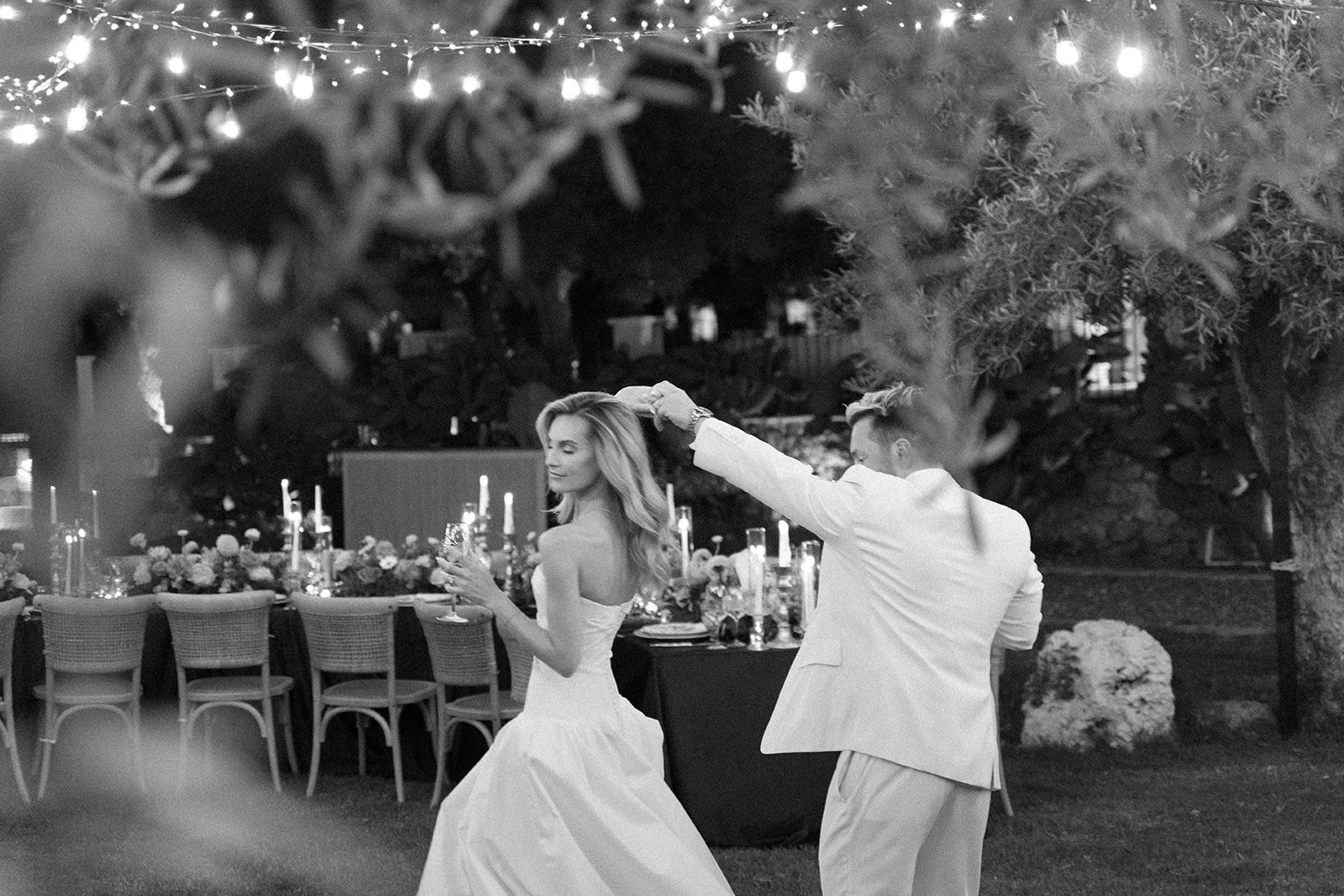 A couple in wedding attire dancing outdoors at night, with string lights overhead and a decorated table in the background.
