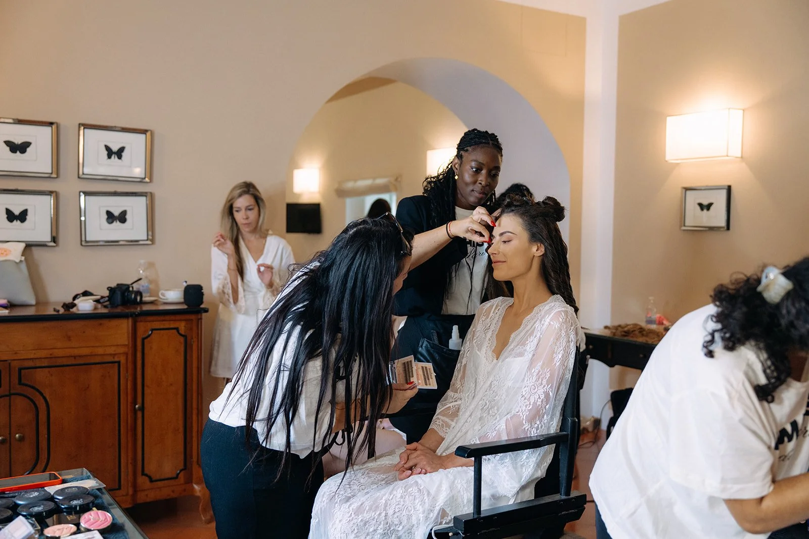 A woman getting her makeup done by a makeup artist at a wedding or special event, with another woman waiting in the background.