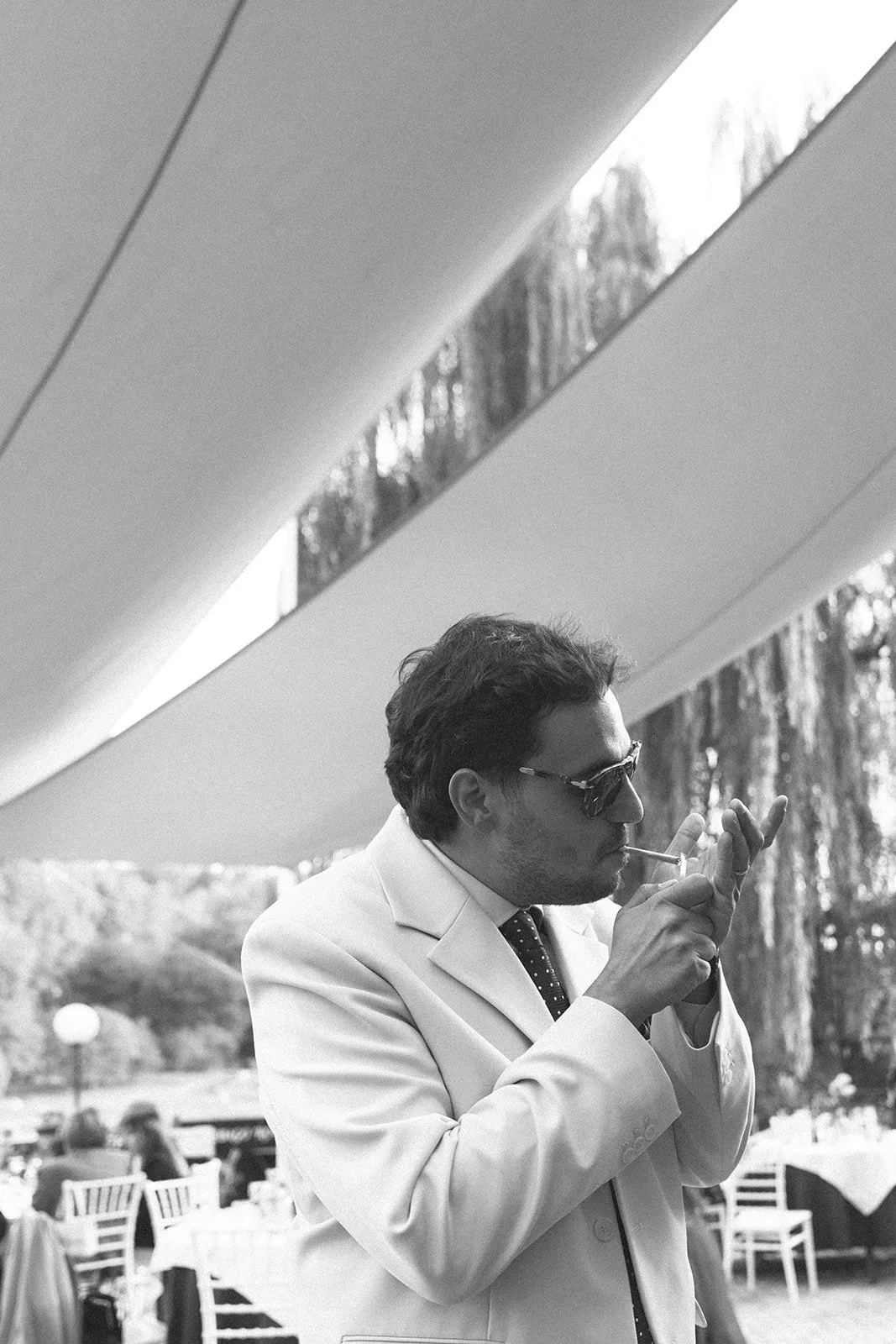 A man with dark hair wearing sunglasses and a white suit jacket, standing outdoors under a modern structure, lighting a cigarette, with trees and outdoor seating in the background.