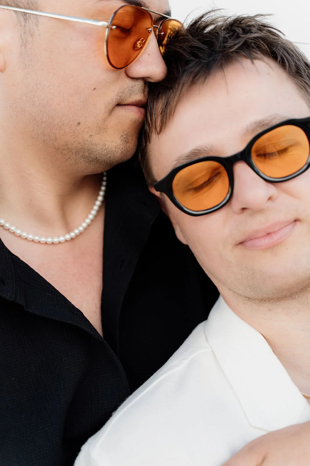 Two men close together, one with a black shirt and pearl necklace, the other in a white shirt, both wearing orange-tinted glasses and with their eyes closed or partially closed.