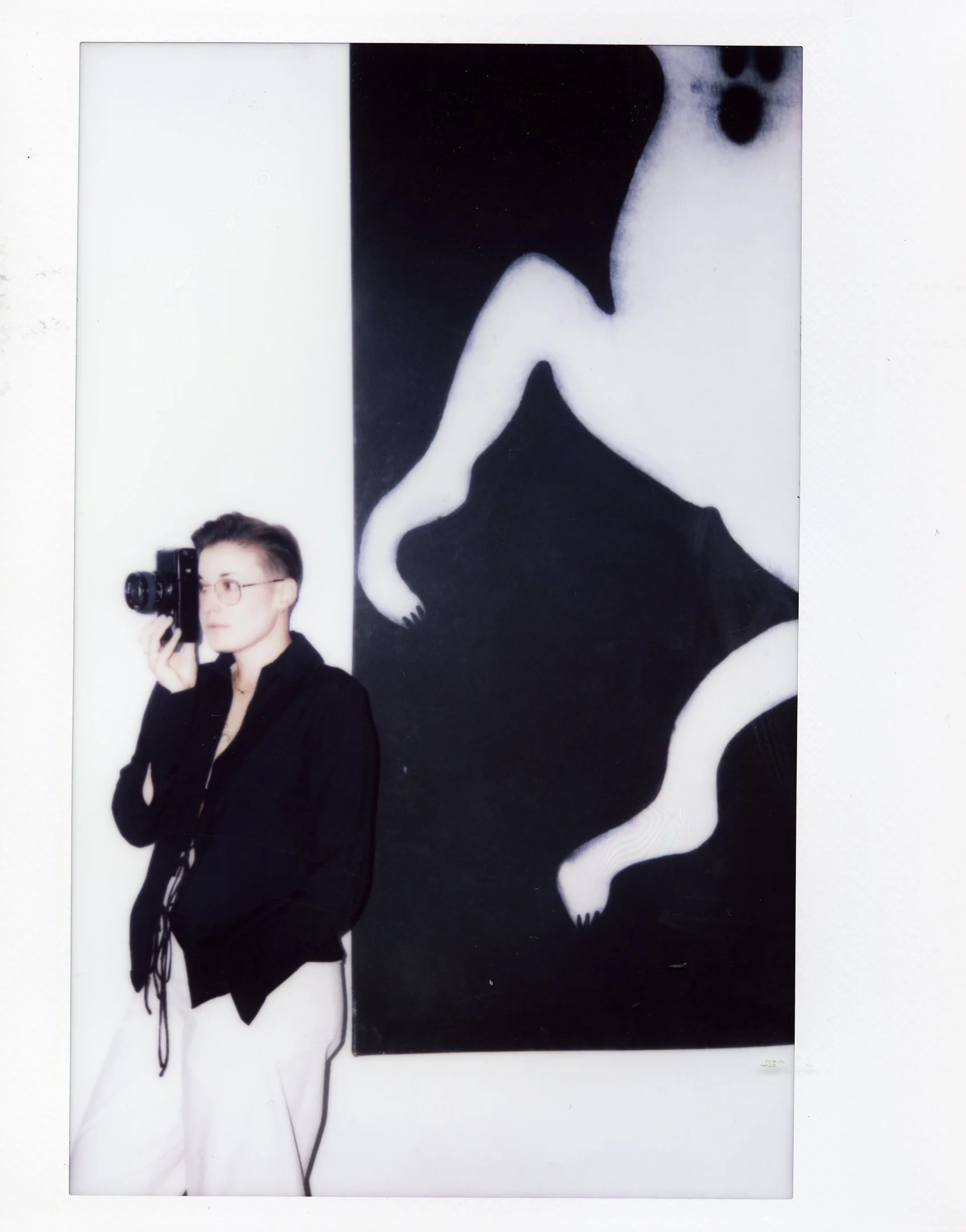 Person with glasses and short hair holding a camera, standing in front of black and white abstract art with animal shapes.