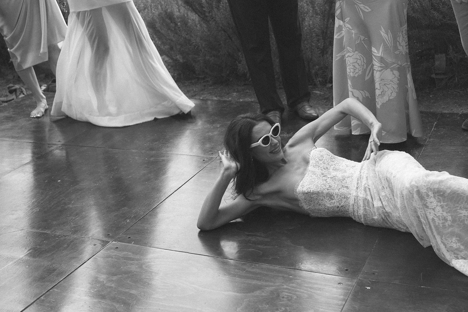 Woman in a lace strapless dress lying on the floor, wearing white sunglasses, smiling, with her left hand bent at the elbow with her hand on her hip. People are standing around her, some in skirts or dresses, in a casual or social setting.