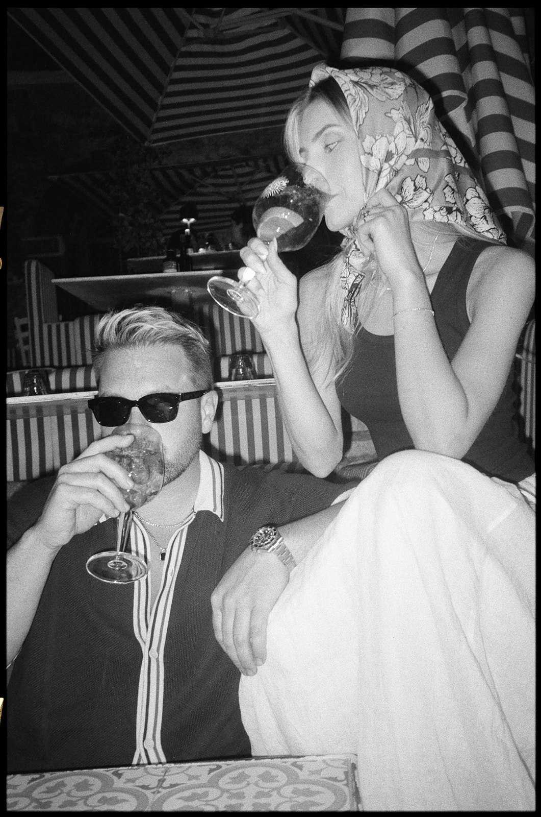 A black-and-white photo of a man and a woman sitting together, both drinking from wine glasses. The man is wearing sunglasses and a striped shirt, while the woman has a floral headscarf, a dark top, and light-colored pants.