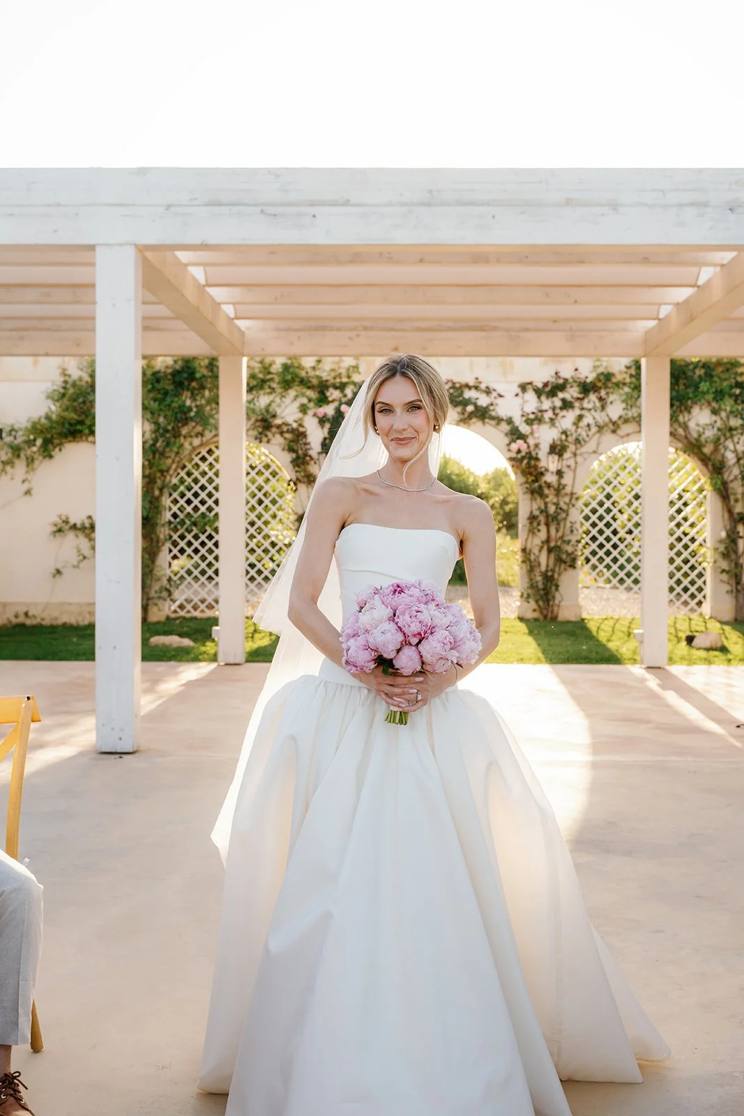 A bride in a white wedding dress holding a pink bouquet, standing outdoors with a pergola and lush greenery in the background.