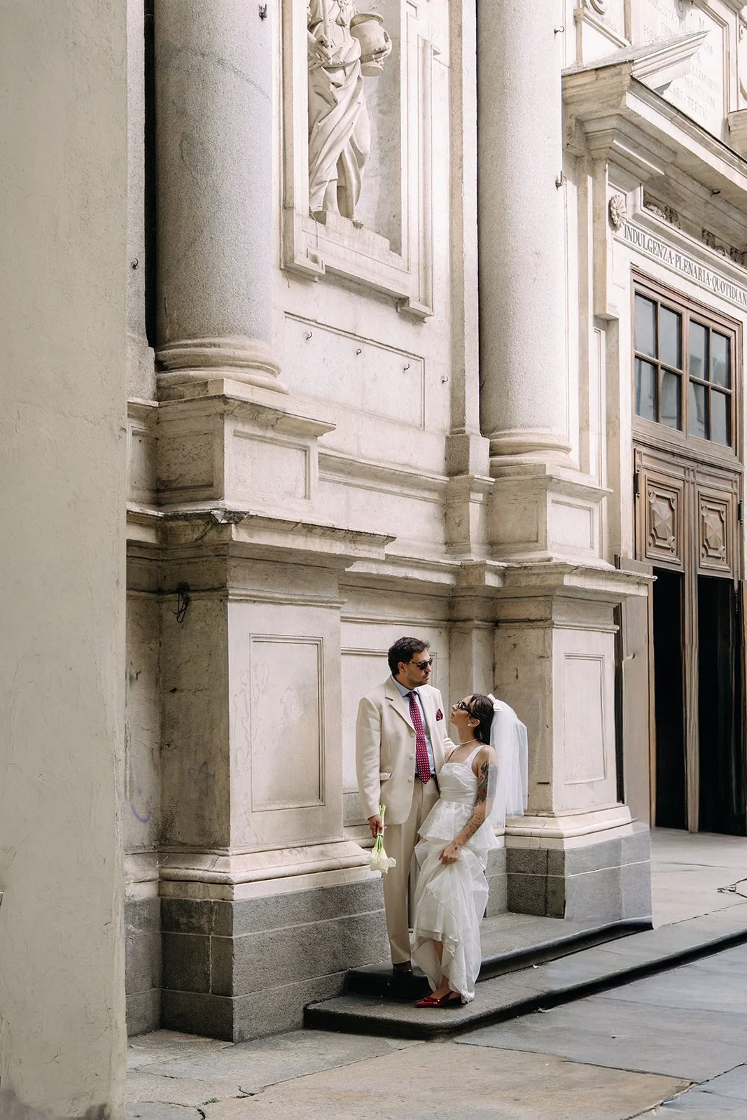 A couple dressed in wedding attire standing together outside a historic building with ornate stone architecture. The groom wears a white suit with a red tie, and the bride wears a white dress with tattoos on her arms, a veil, and red shoes, holding a