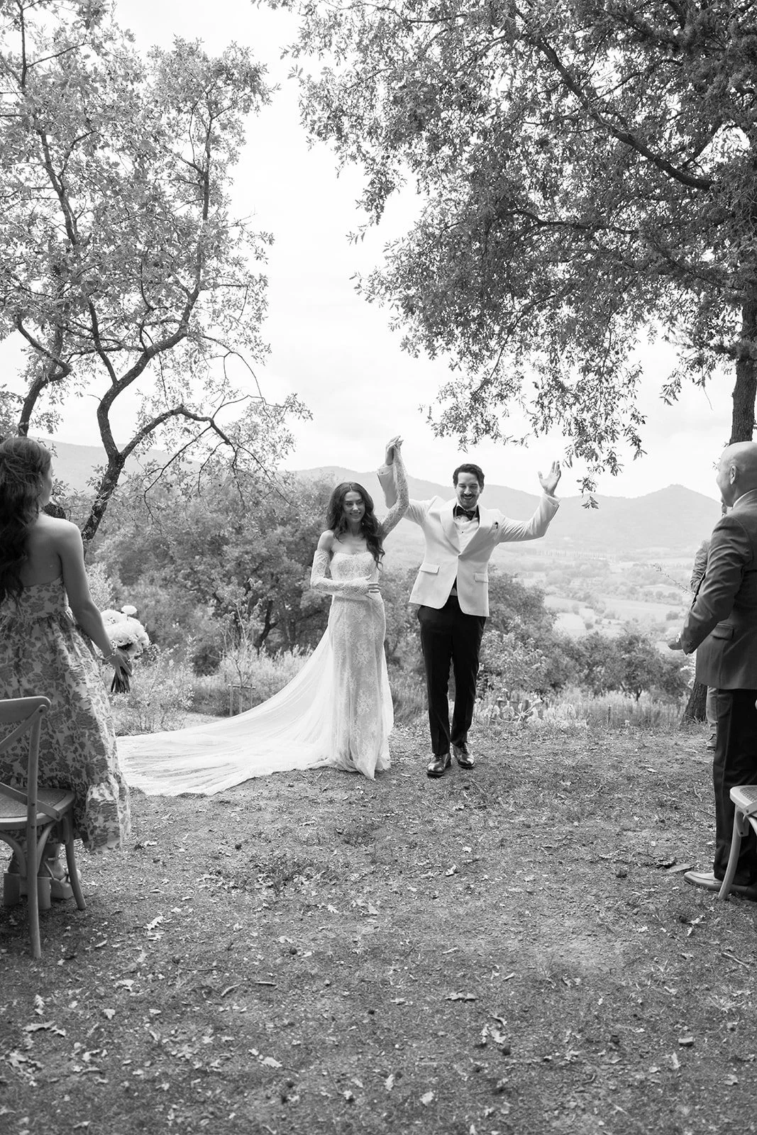 A black and white photo of a wedding ceremony outdoors, with the bride and groom holding hands and raising them in celebration. The bride is in a long, elegant wedding dress with lace details, and the groom is in a white tuxedo jacket with dark pants