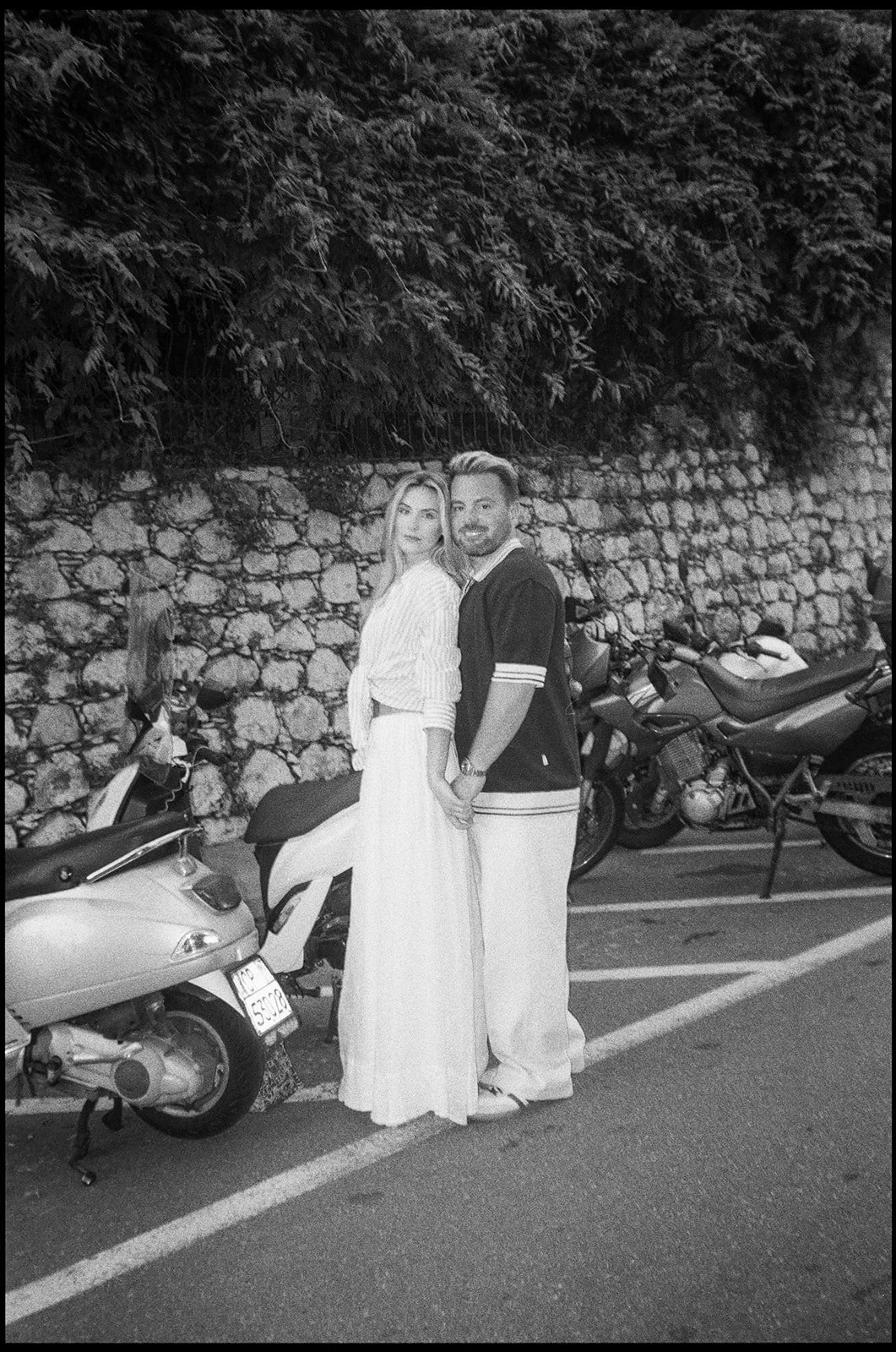 A couple holding hands standing next to parked scooters on a street at night, with a stone wall and greenery in the background.