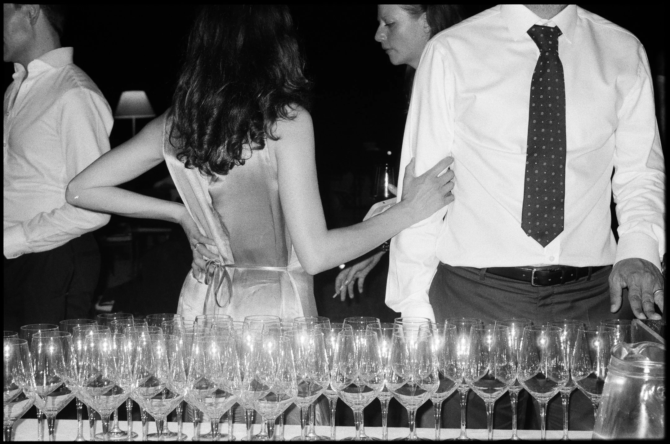 Black and white photo of a woman and a man at a party, with a table full of empty wine glasses in front of them. The woman has long curly hair and is wearing a sleeveless dress. The man is wearing a white shirt and a dark tie with polka dots.