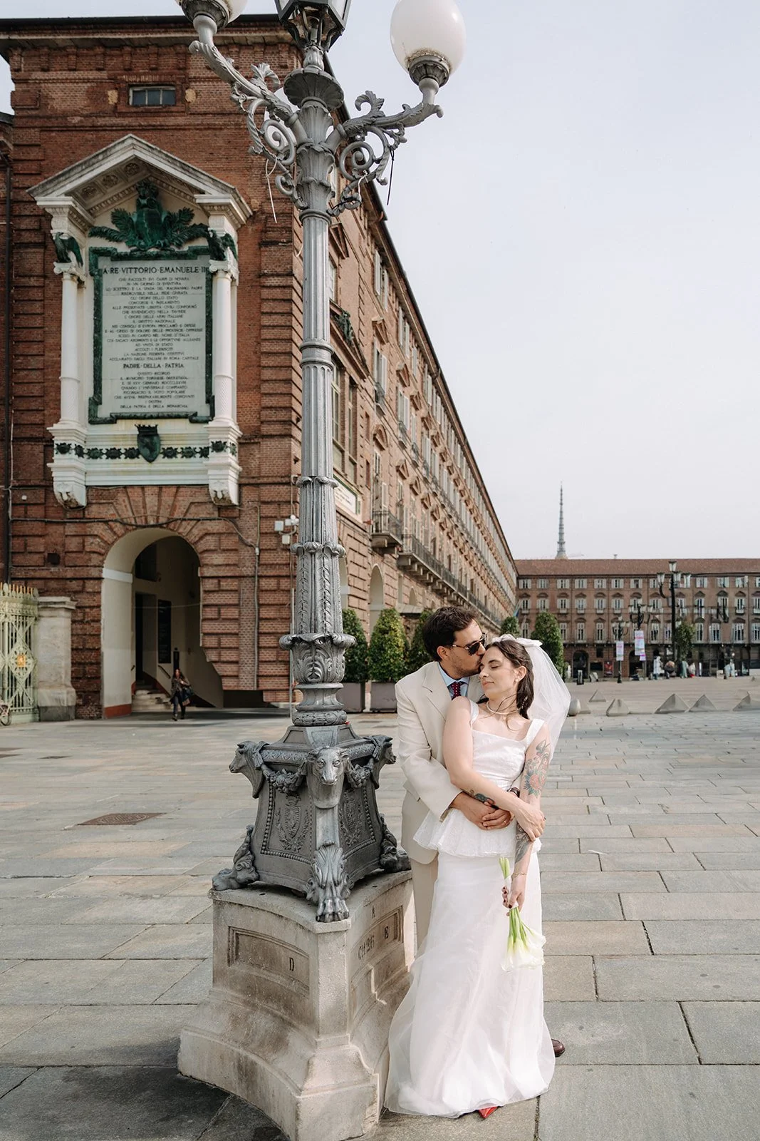 A newlywed couple sharing a kiss on a city square, with the groom in a white suit and the bride in a white wedding dress holding flowers, standing beside an ornate street lamp with a historic building in the background.