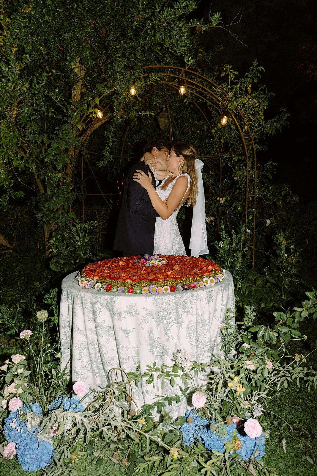 A couple kissing during a wedding celebration, standing behind a heart-shaped arrangement of strawberries and flowers on a table, surrounded by greenery at night.