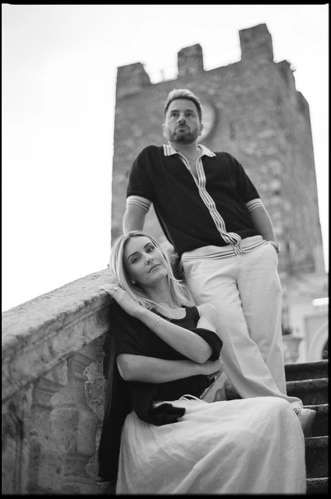 A black and white photo of a man and a woman outdoors. The woman is sitting on stone stairs, resting her head on her hand, looking at the camera. The man is standing behind her, leaning against the stone railing, looking away. Tall stone tower is in 