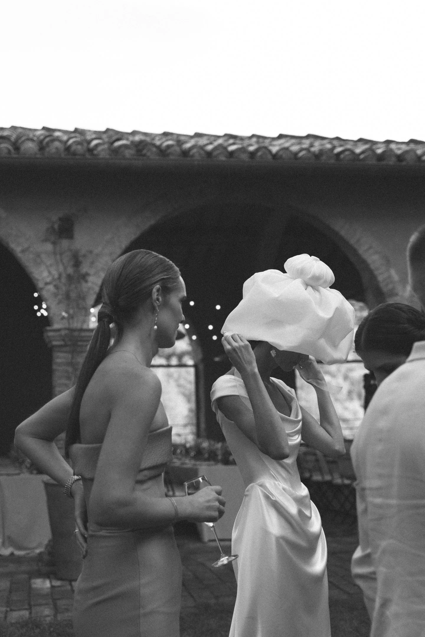 Group of people at an outdoor event in the evening, with a woman in a strapless dress holding a glass of wine, another woman adjusting a large, elaborate hat, and a man wearing a button-down shirt.