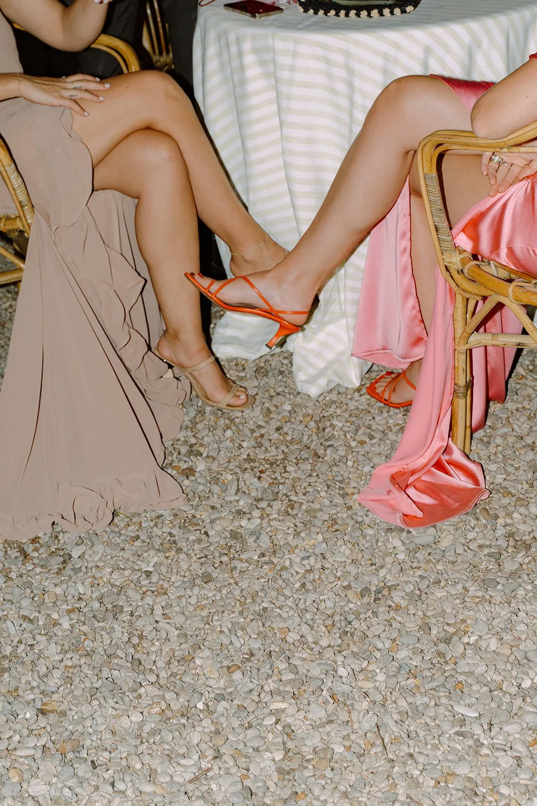 Close-up of two women sitting on wicker chairs with their legs extended, showing their footwear and dresses. One woman is wearing beige high heels and a beige dress, and the other is wearing orange high heels and a pink satin dress with a slit.