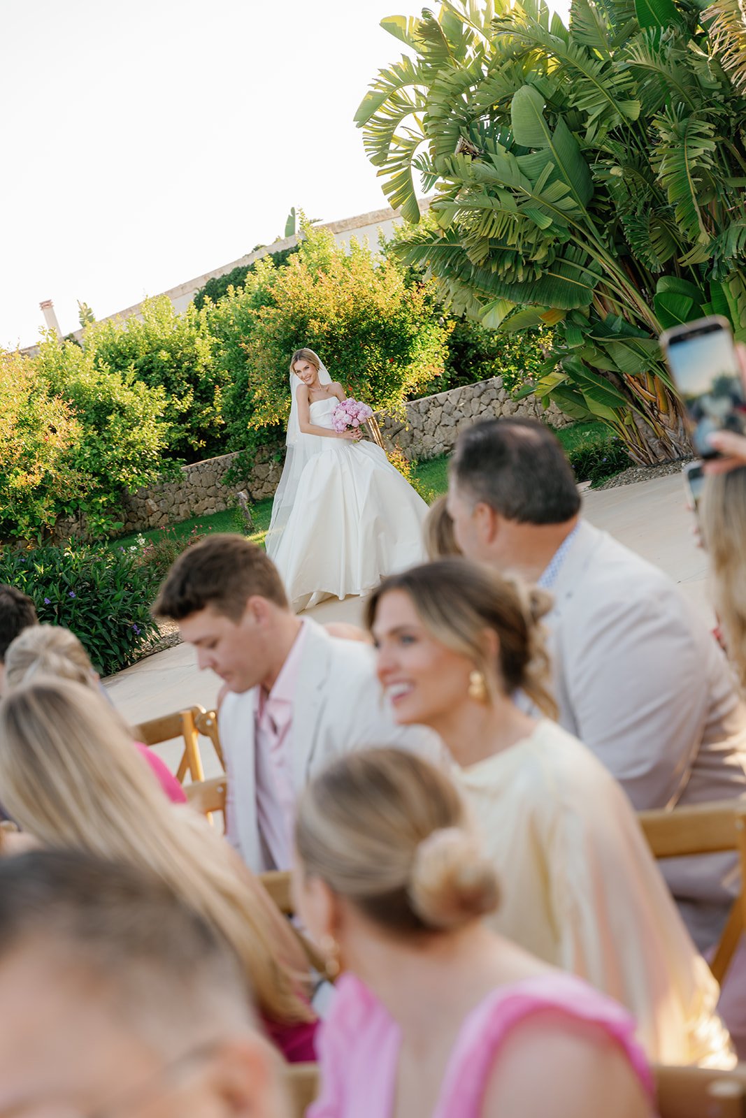 A bride in a white wedding dress standing outside, holding a pink bouquet, during a wedding ceremony with guests seated nearby, lush green trees and a stone wall in the background, and sunlight illuminating the scene.