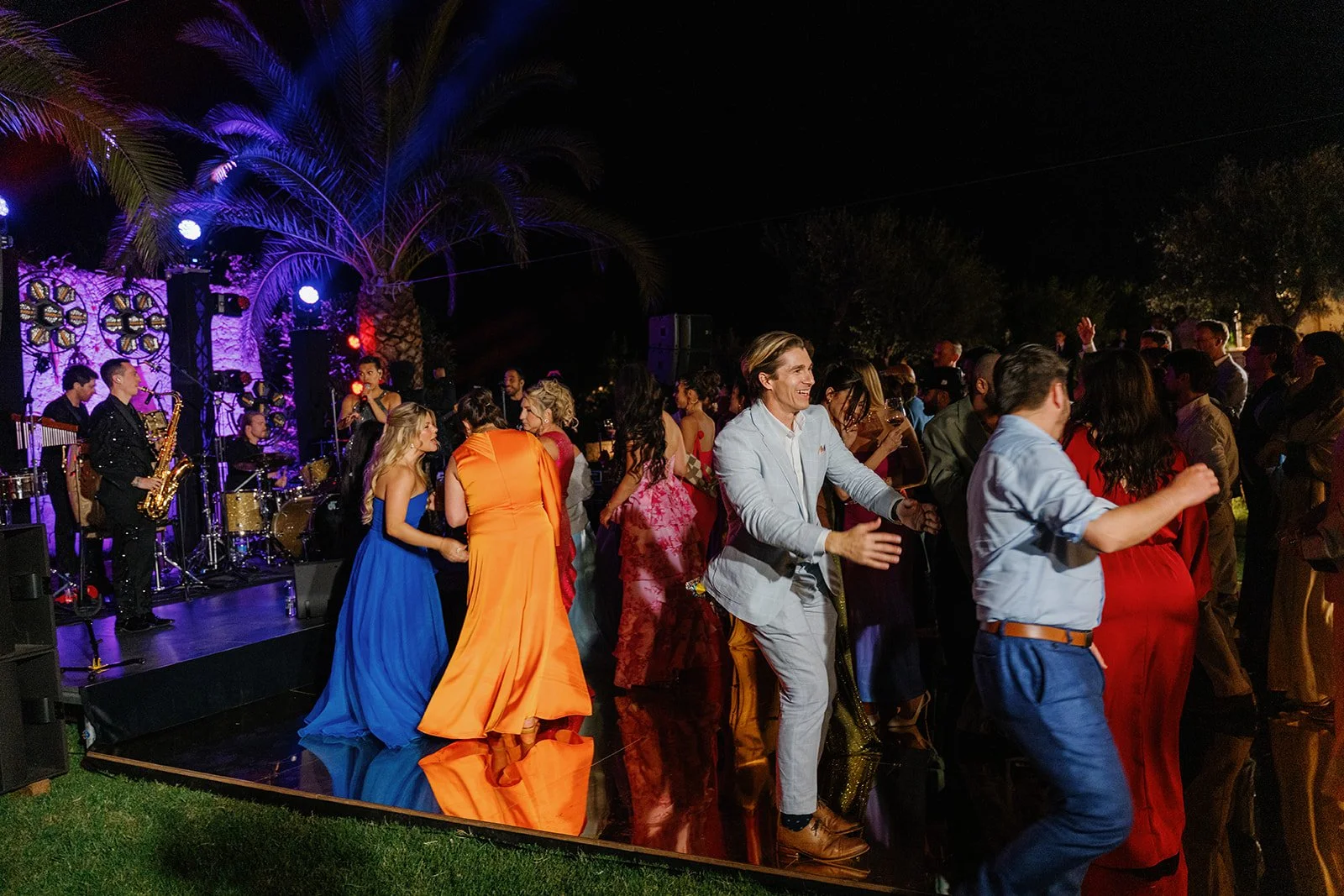 People dancing and enjoying live music at an outdoor night event with a band playing on stage and colorful lighting.