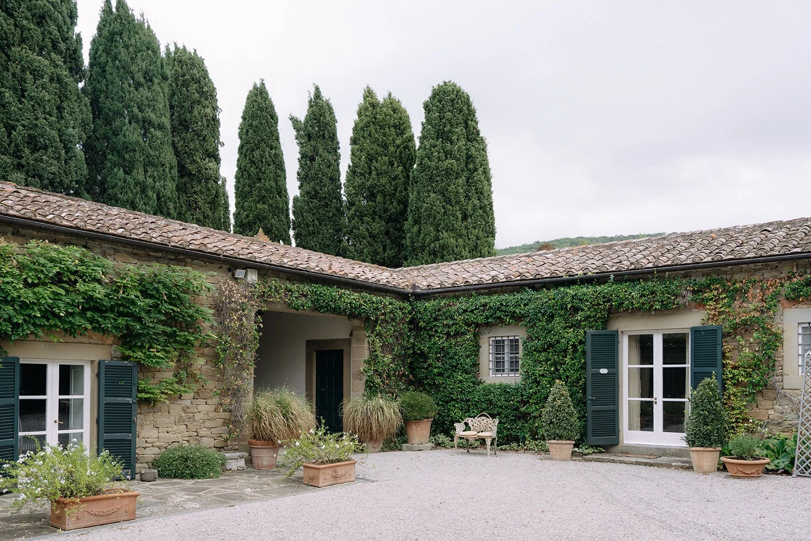 A rustic stone house with dark green shutters, surrounded by potted plants and greenery, with large trees in the background under a cloudy sky.