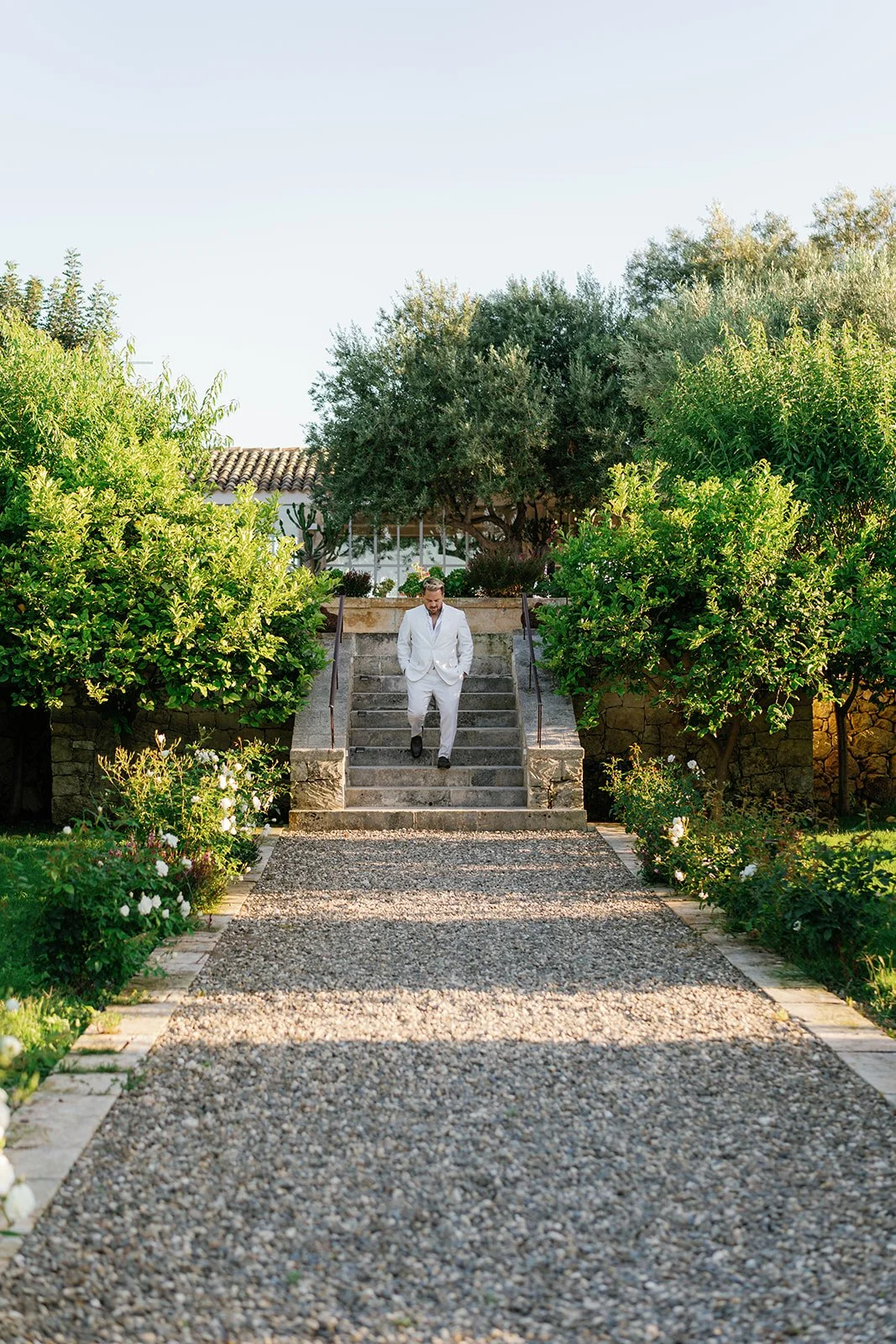 A man in a white suit walking down stone steps in a lush garden with green trees and bushes, under a clear blue sky.
