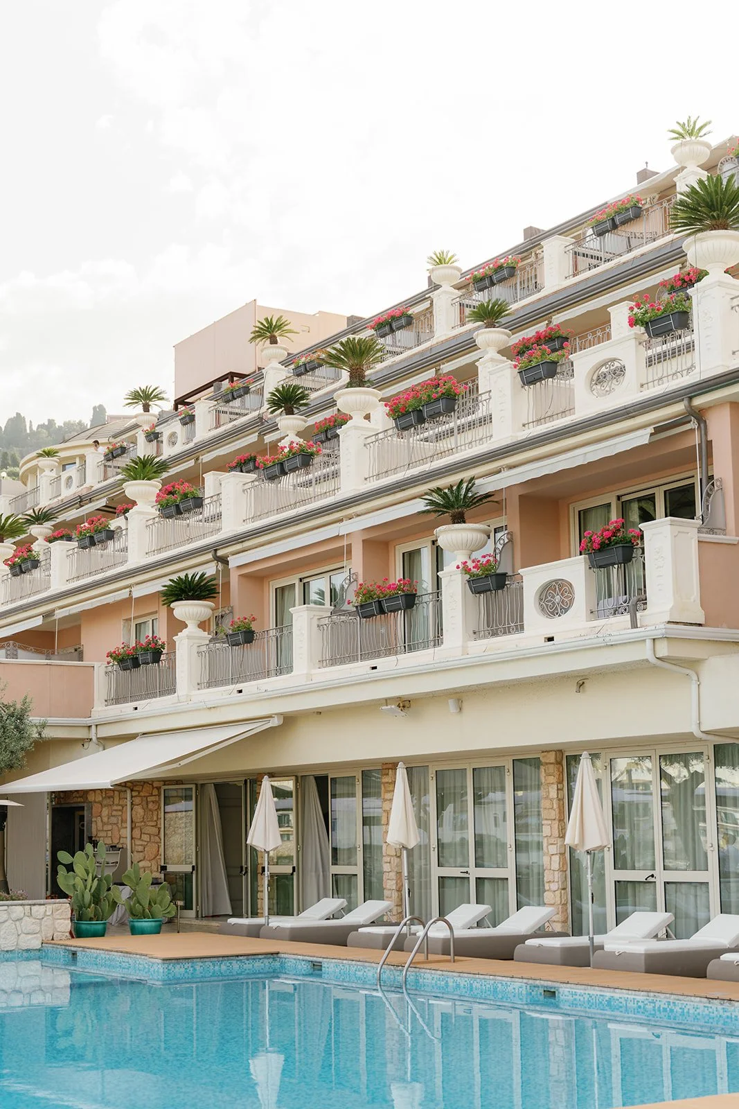 A hotel with a swimming pool in front, multiple balconies with potted plants, pink flowers, and umbrellas.