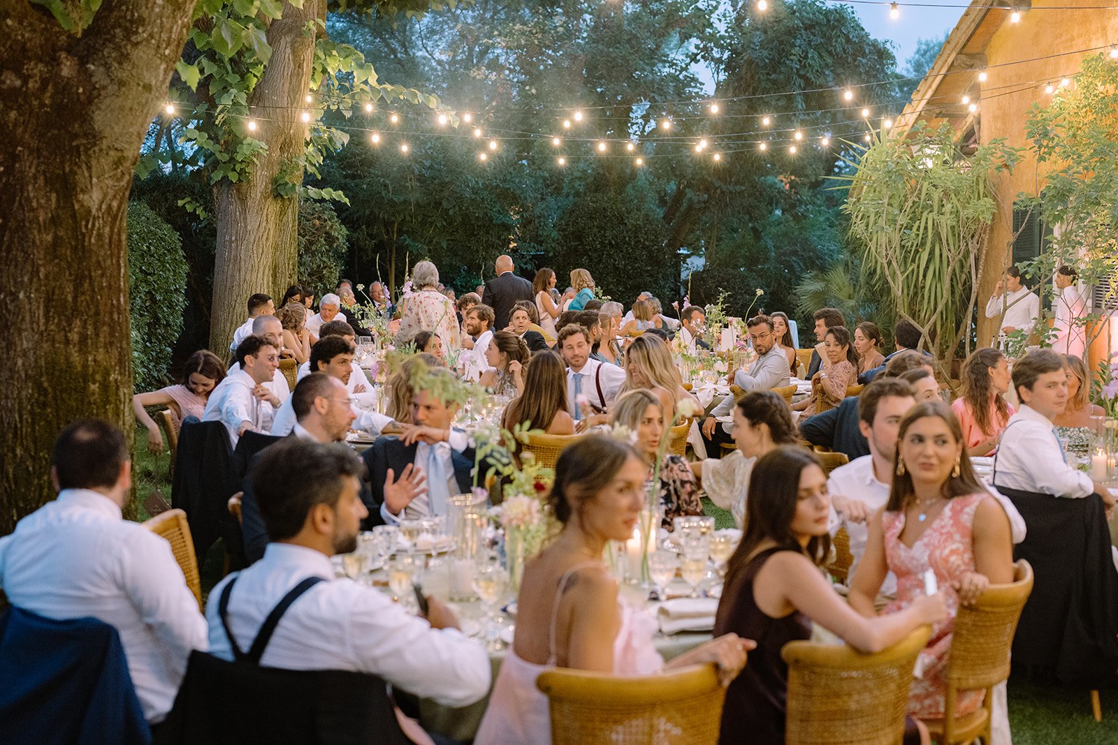 An outdoor evening gathering with many people seated at long tables decorated with flowers and candles, under string lights, surrounded by trees and greenery.