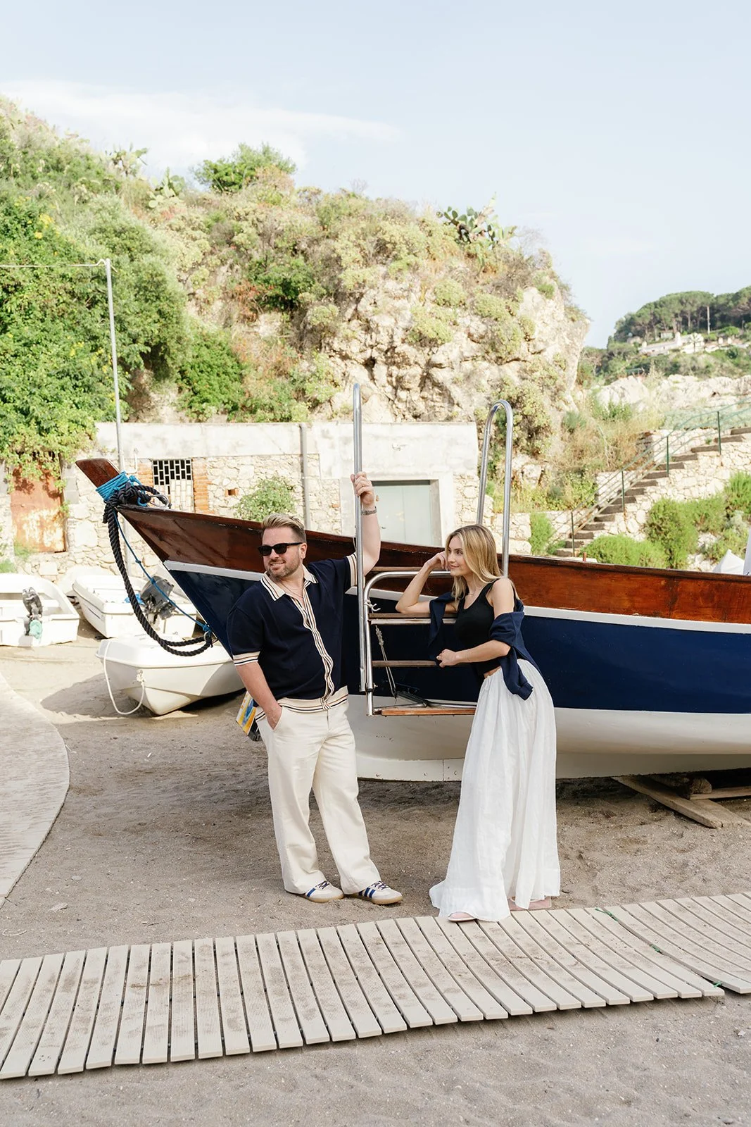 A man and woman standing next to a boat at a marina, with the man holding onto a metal ladder and the woman leaning on it, surrounded by boats and rocky hills in the background.
