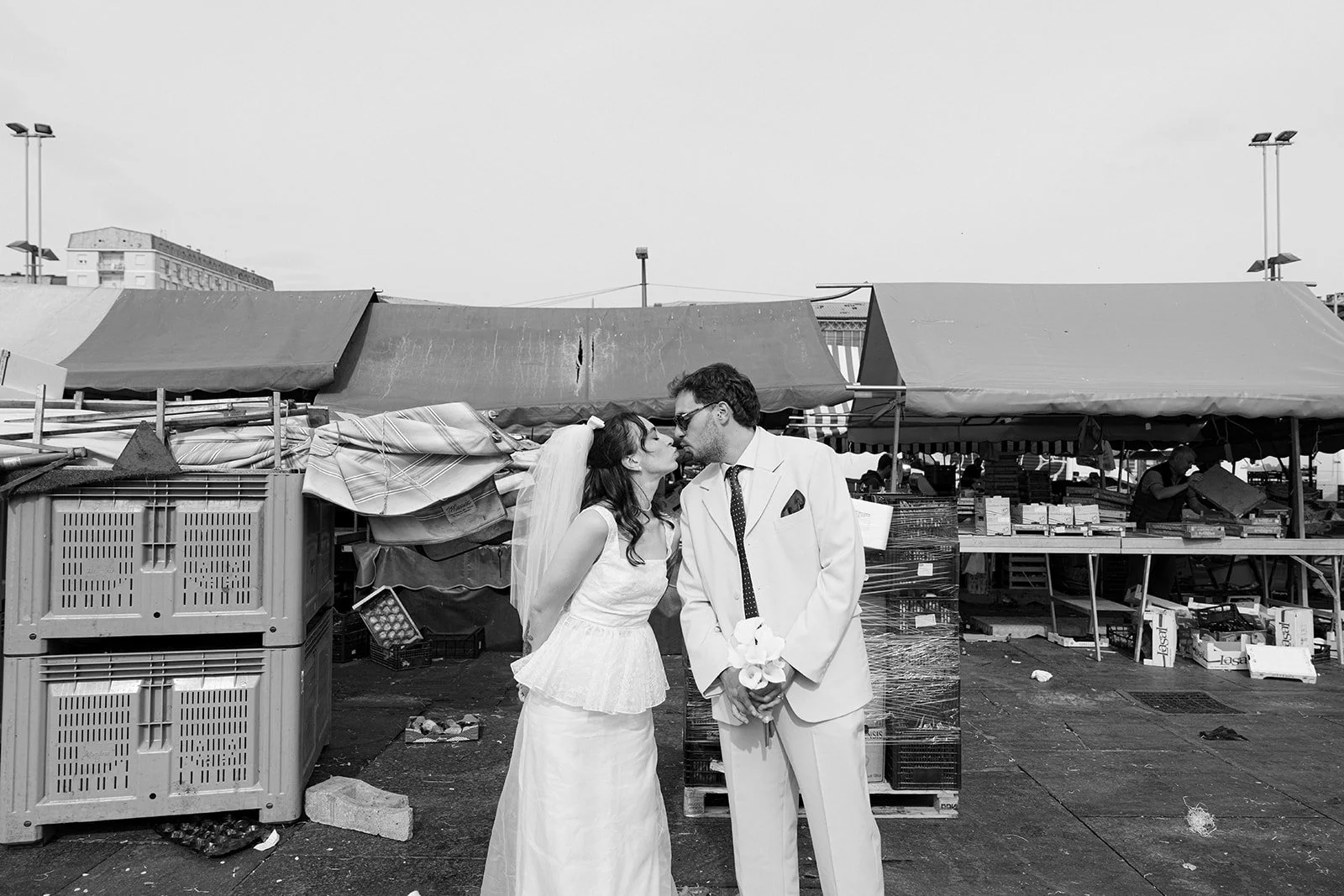 Black and white photo of a couple in wedding attire sharing a kiss in front of an outdoor market with stalls and people in the background.