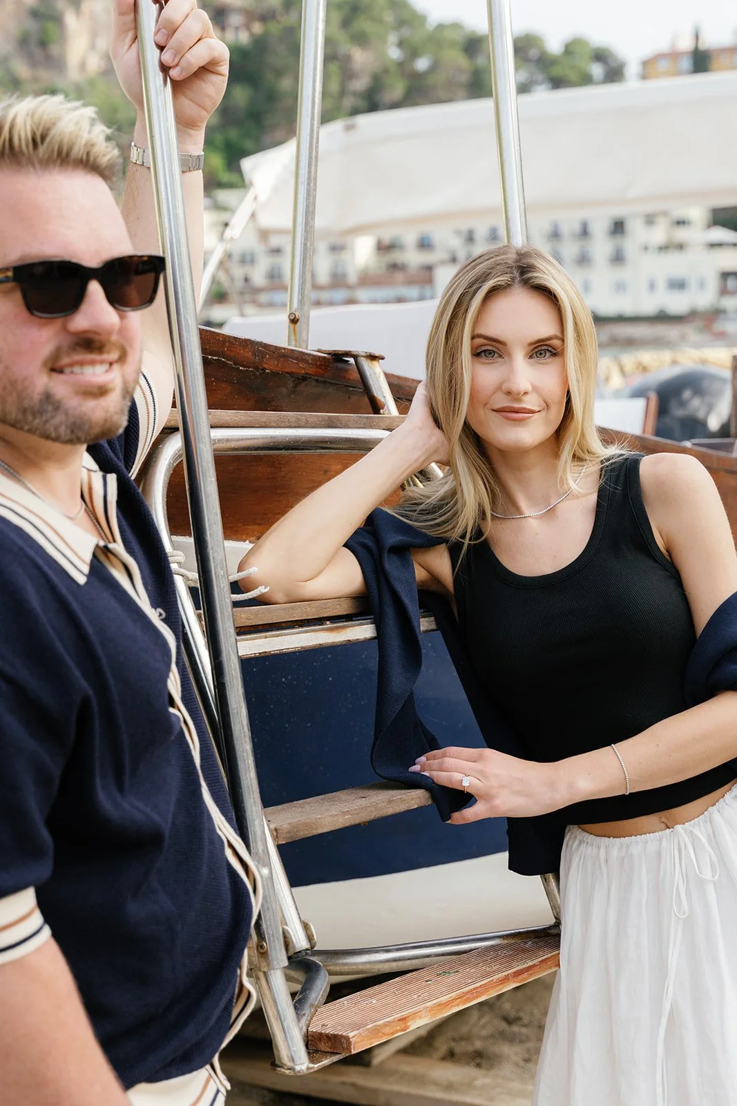 A woman with blonde hair, wearing a black sleeveless top and white skirt, leaning against a docked boat, and a man with blonde hair, sunglasses, and a navy striped polo shirt, standing nearby.