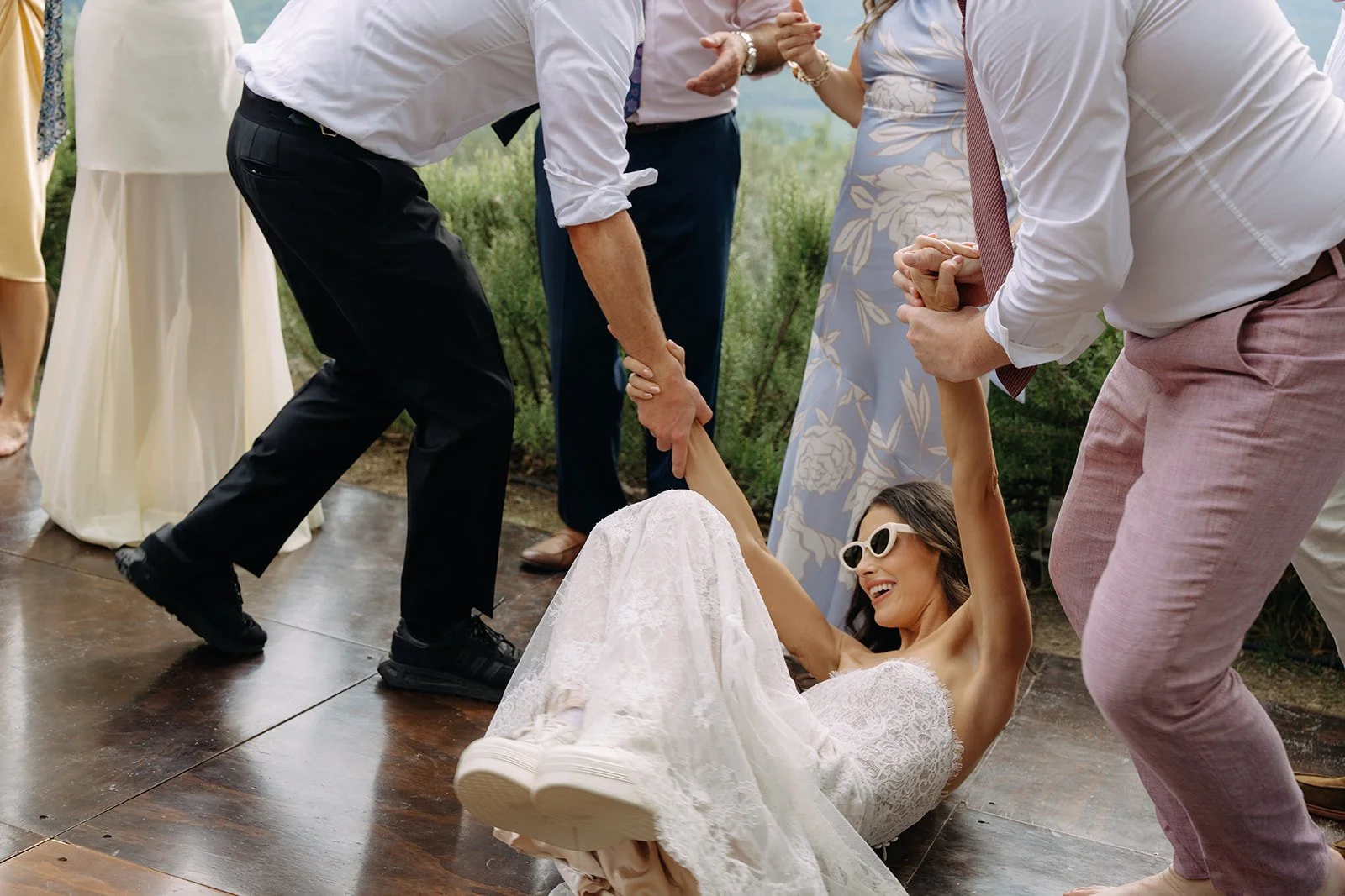 A woman in a wedding dress and sunglasses sitting on the floor, smiling as she is handcuffed by a man, while others around her are involved in the playful activity at an outdoor wedding reception.