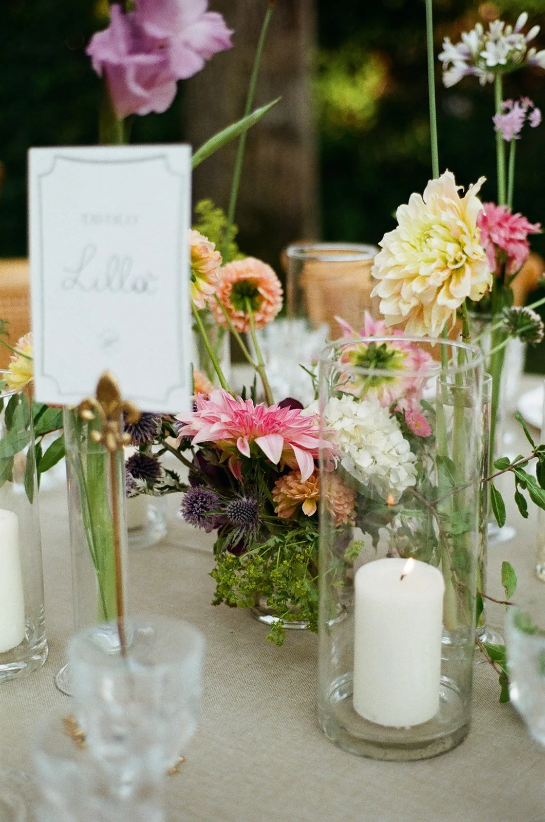 A floral arrangement with various pink, cream, and purple flowers in glass vases, with a lit white candle inside a tall glass holder, and a small table sign that reads 'Table Lella', on a beige tablecloth.